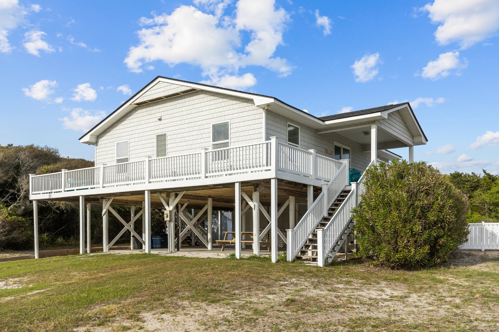 Elevated beach house with wraparound deck and stairs, surrounded by coastal vegetation under blue sky.