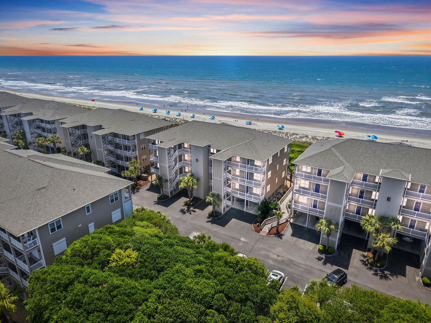 Beachfront property complex with direct ocean access, sandy shoreline, and colorful umbrellas dotting the coast during golden hour.