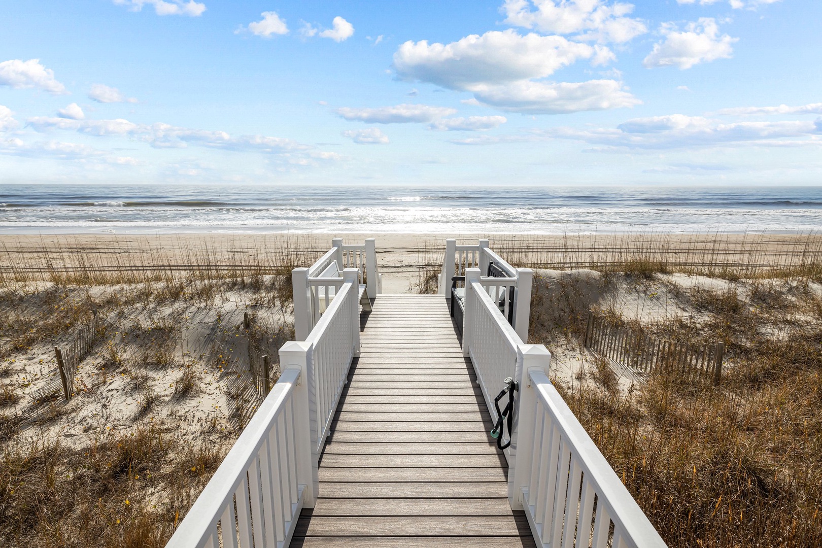 A wooden boardwalk leads through coastal dunes to pristine sandy shores where ocean waves meet endless blue skies.