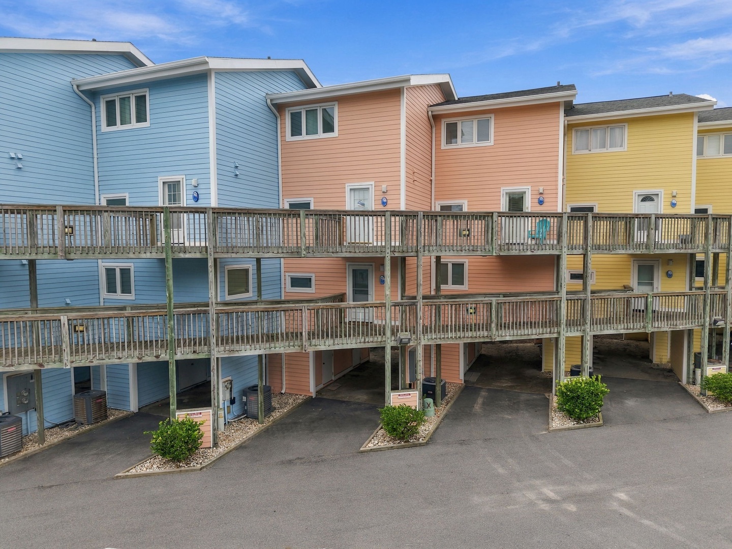 Colorful row of coastal vacation homes with multiple levels and parking beneath each unit, surrounded by a welcoming neighborhood setting.
