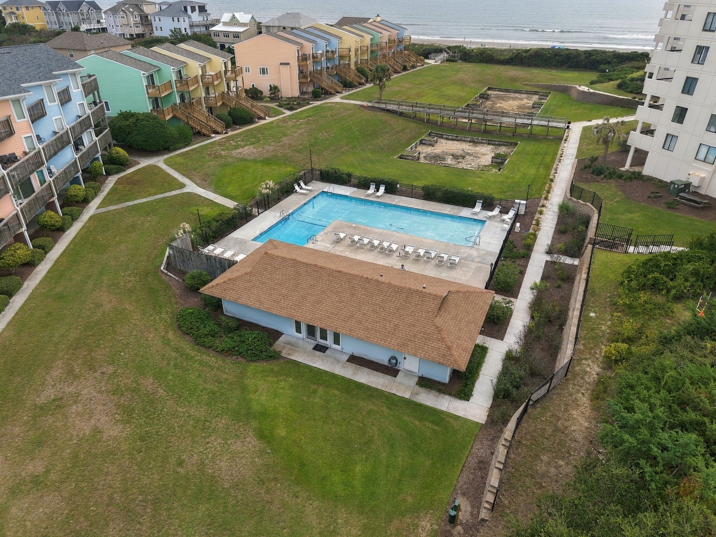 Aerial view of the beachfront property featuring a sparkling pool, communal lawn areas, and boardwalk access to the sandy shore. The oceanfront complex includes colorful buildings and a playground area for guests.