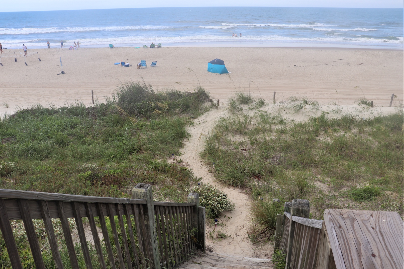 A wooden walkway leads through coastal dunes to the expansive sandy beach where gentle waves meet the shore.