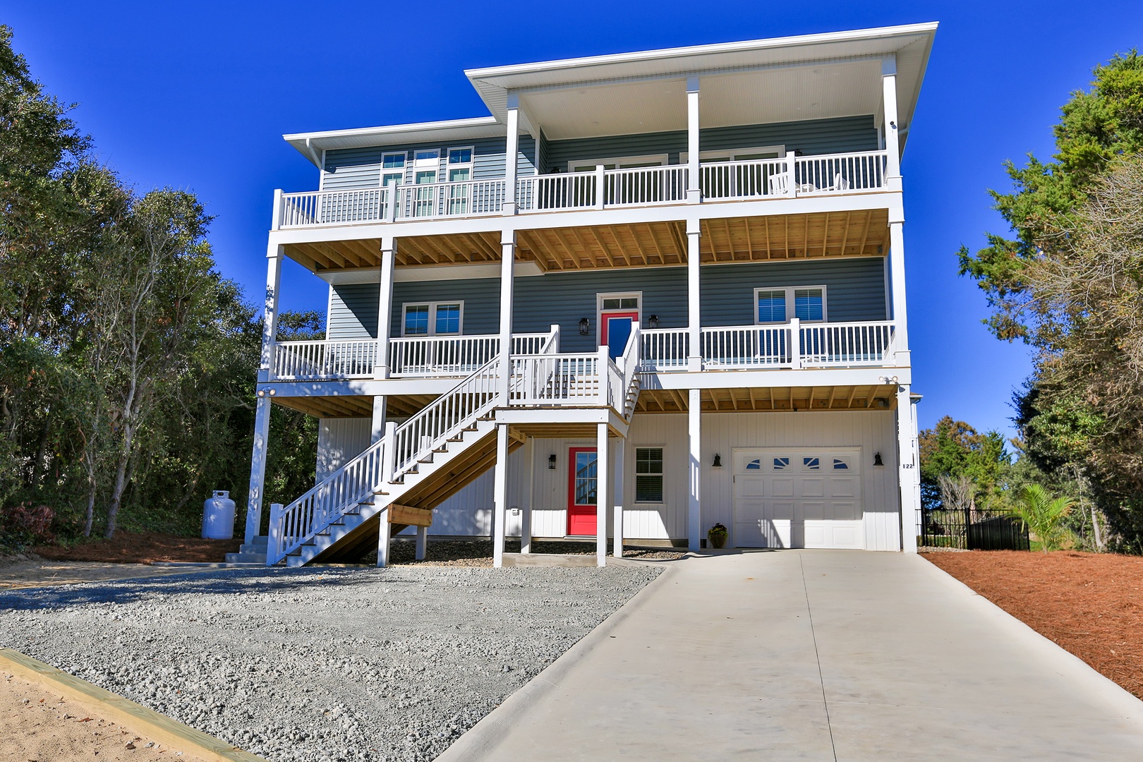 Modern three-story coastal home with spacious balconies, garage parking, and inviting blue exterior surrounded by natural landscaping.