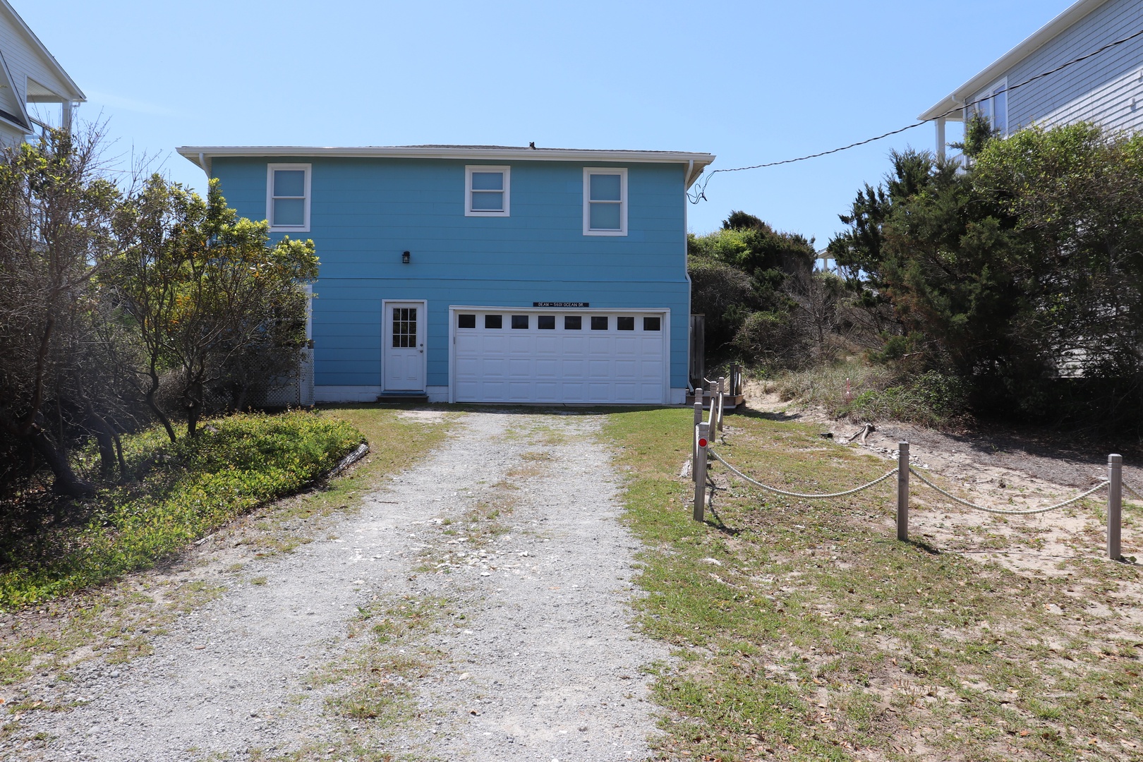 Charming blue coastal home with private driveway and garage, nestled among natural vegetation in a peaceful neighborhood setting.