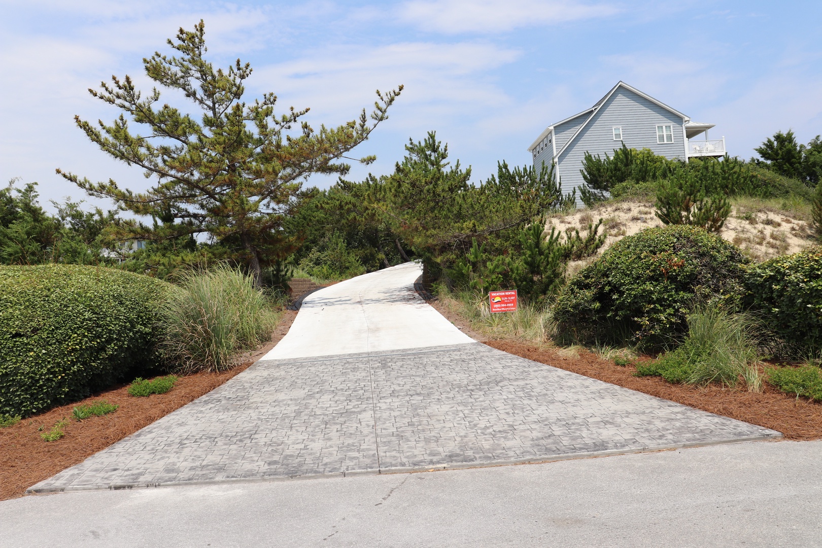 A paved driveway leads through coastal landscaping to a charming gray beach house nestled among native plants and dunes.