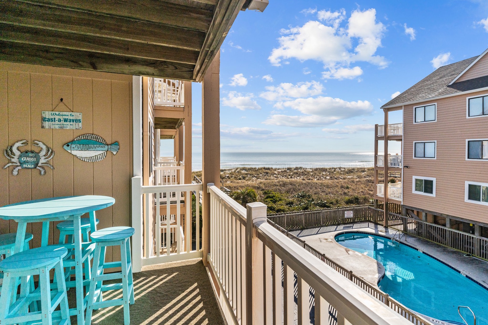 Your private balcony overlooks the sparkling pool and ocean beyond, where turquoise bar stools invite morning coffee with coastal views.