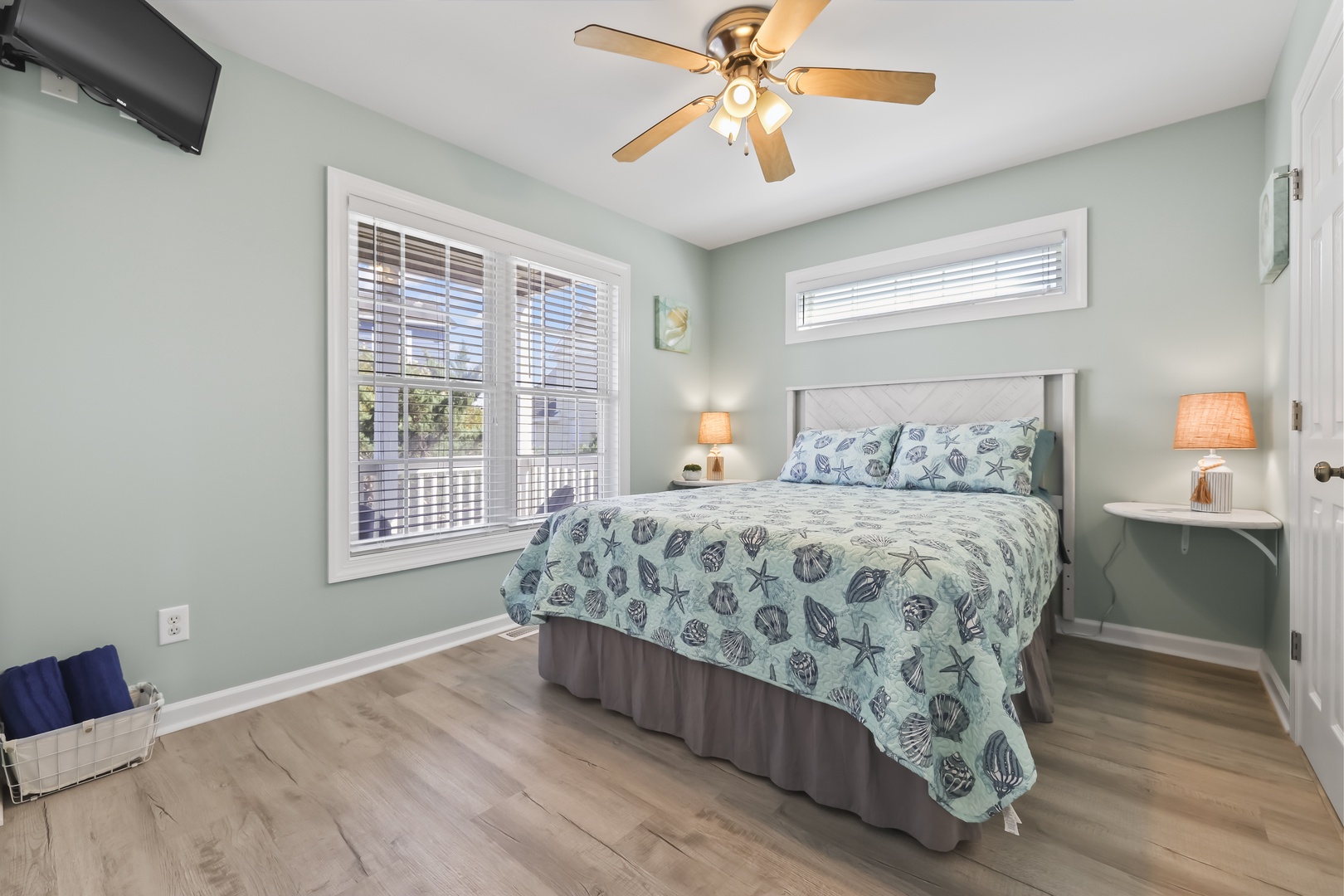 Relax in your serene bedroom with coastal charm, featuring a queen bed dressed in soft ocean-themed linens, ceiling fan, TV, warm lighting, and peaceful views through bright windows.