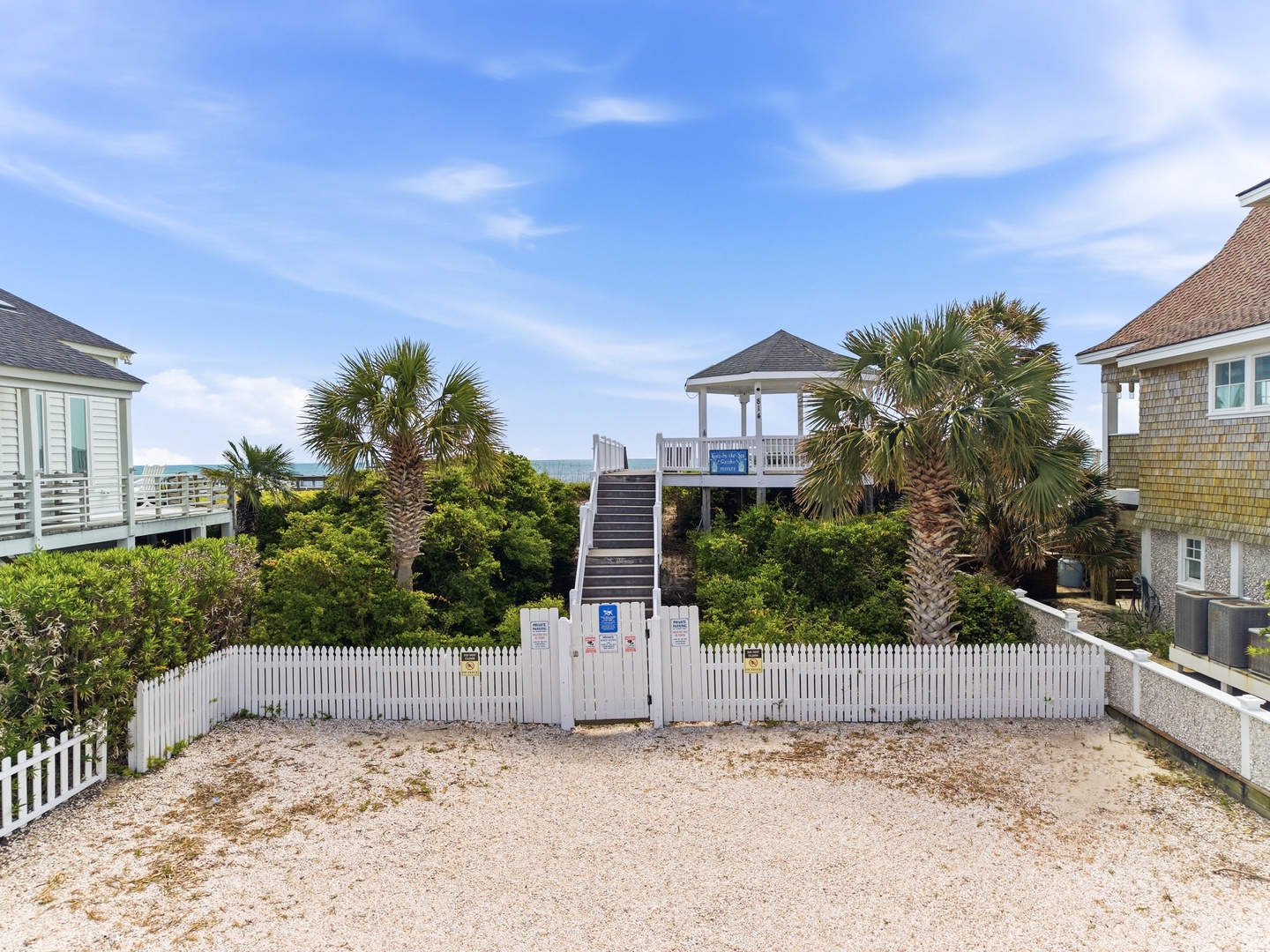 Beach access boardwalk flanked by palm trees and coastal homes, connecting the property to sandy shores under clear blue skies.