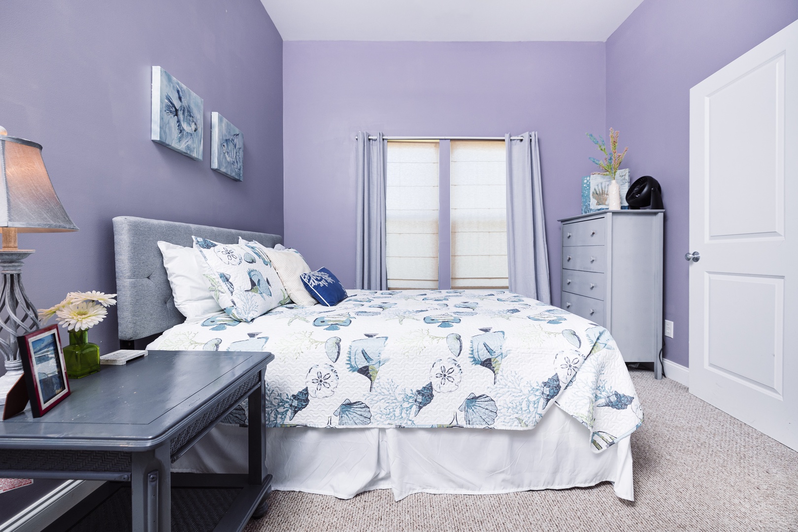 Drift off in this serene lavender bedroom with soft blue accents, cozy bedding, and peaceful ocean-themed artwork.