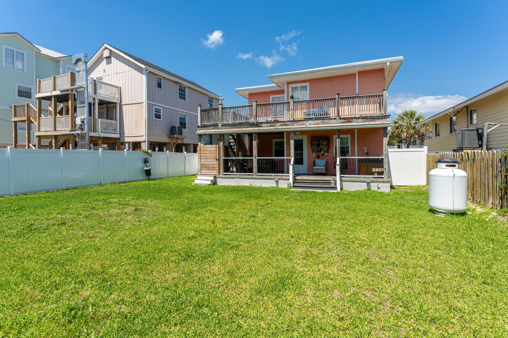 Coral-pink coastal retreat with spacious deck overlooks a grassy backyard, surrounded by neighboring beach homes under a bright blue sky—ideal for outdoor gatherings and relaxing coastal living.