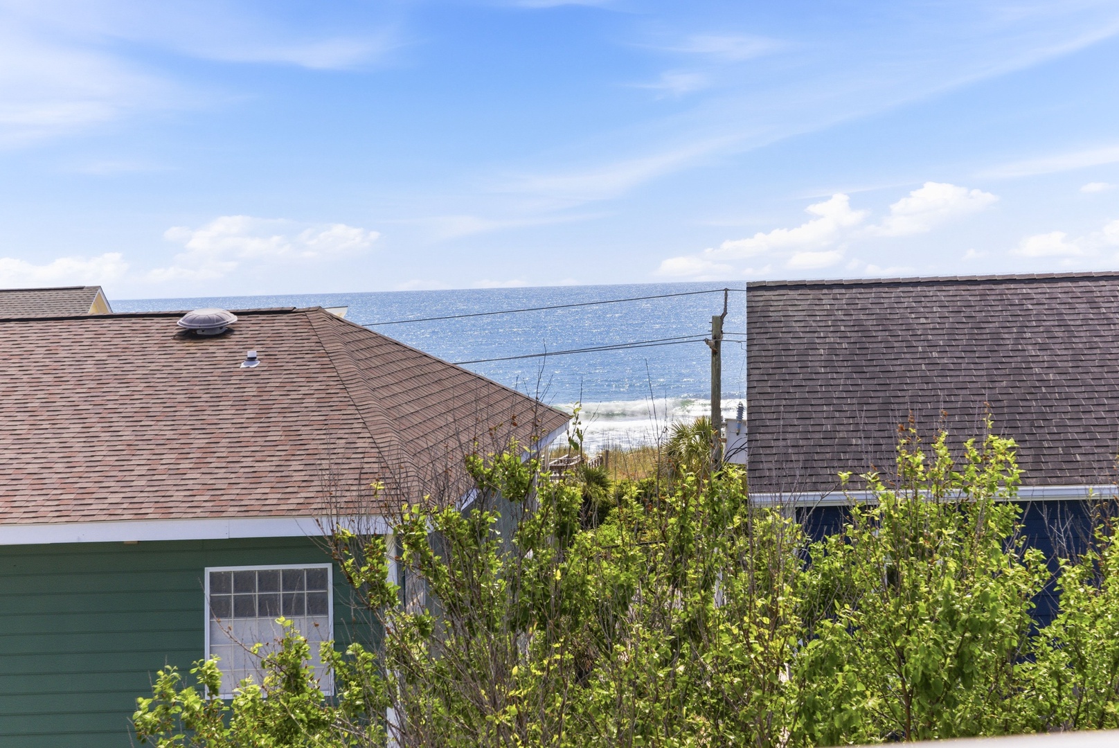 Coastal property nestled among neighboring homes with ocean views visible beyond rooftops and lush green foliage marking the entrance to the area.
