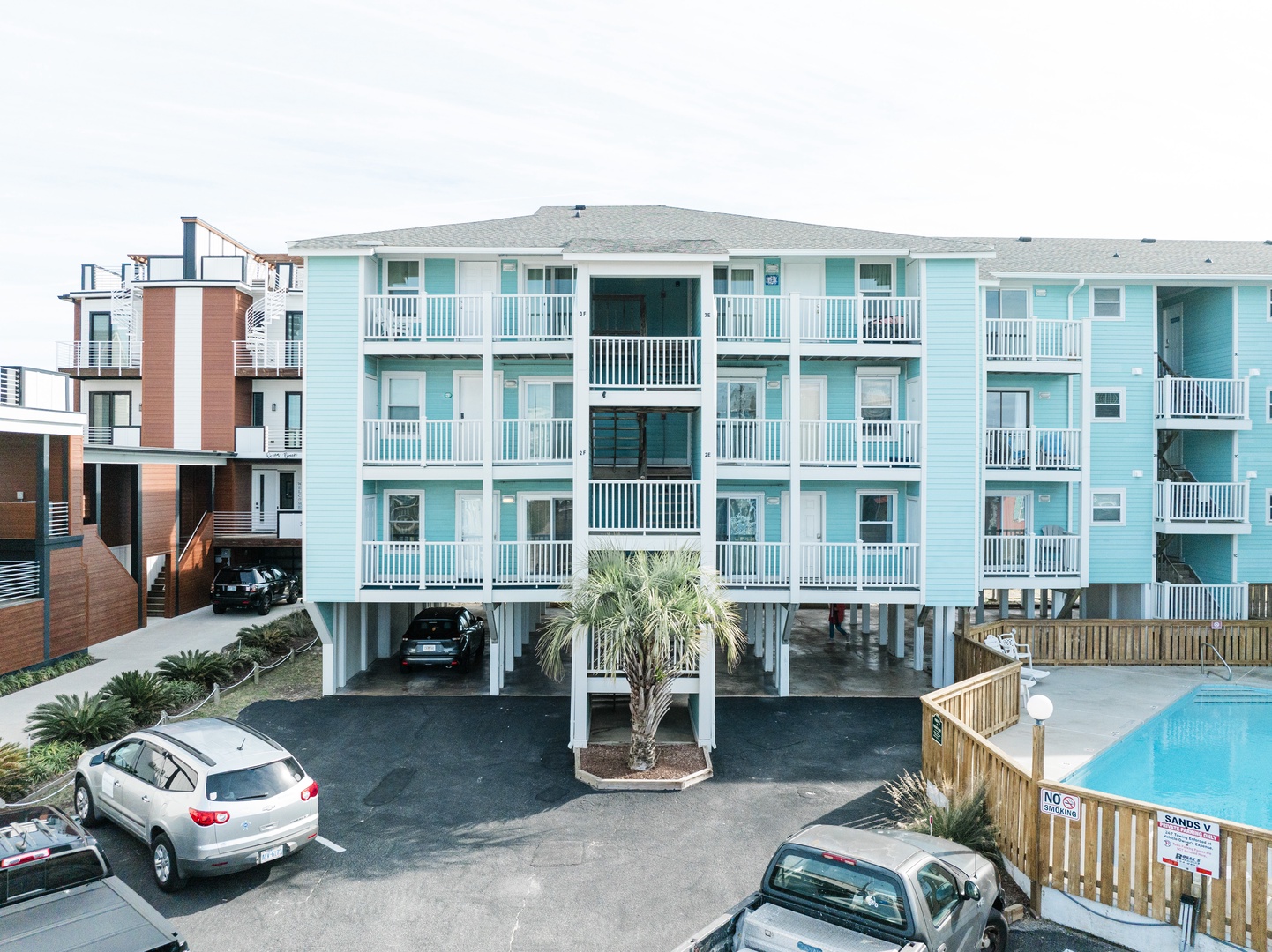 Coastal rental complex with bright turquoise units, private balconies, covered parking, and an inviting pool area surrounded by tropical landscaping.