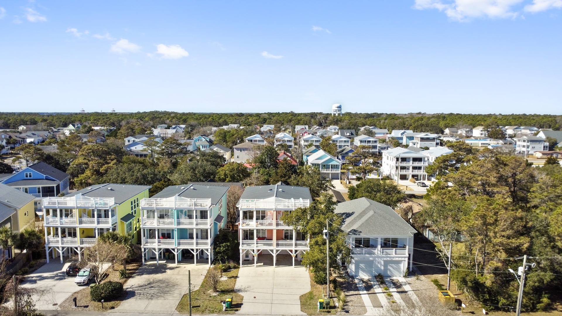 Aerial view of a charming coastal neighborhood featuring colorful beach houses nestled among mature trees under a bright blue sky.