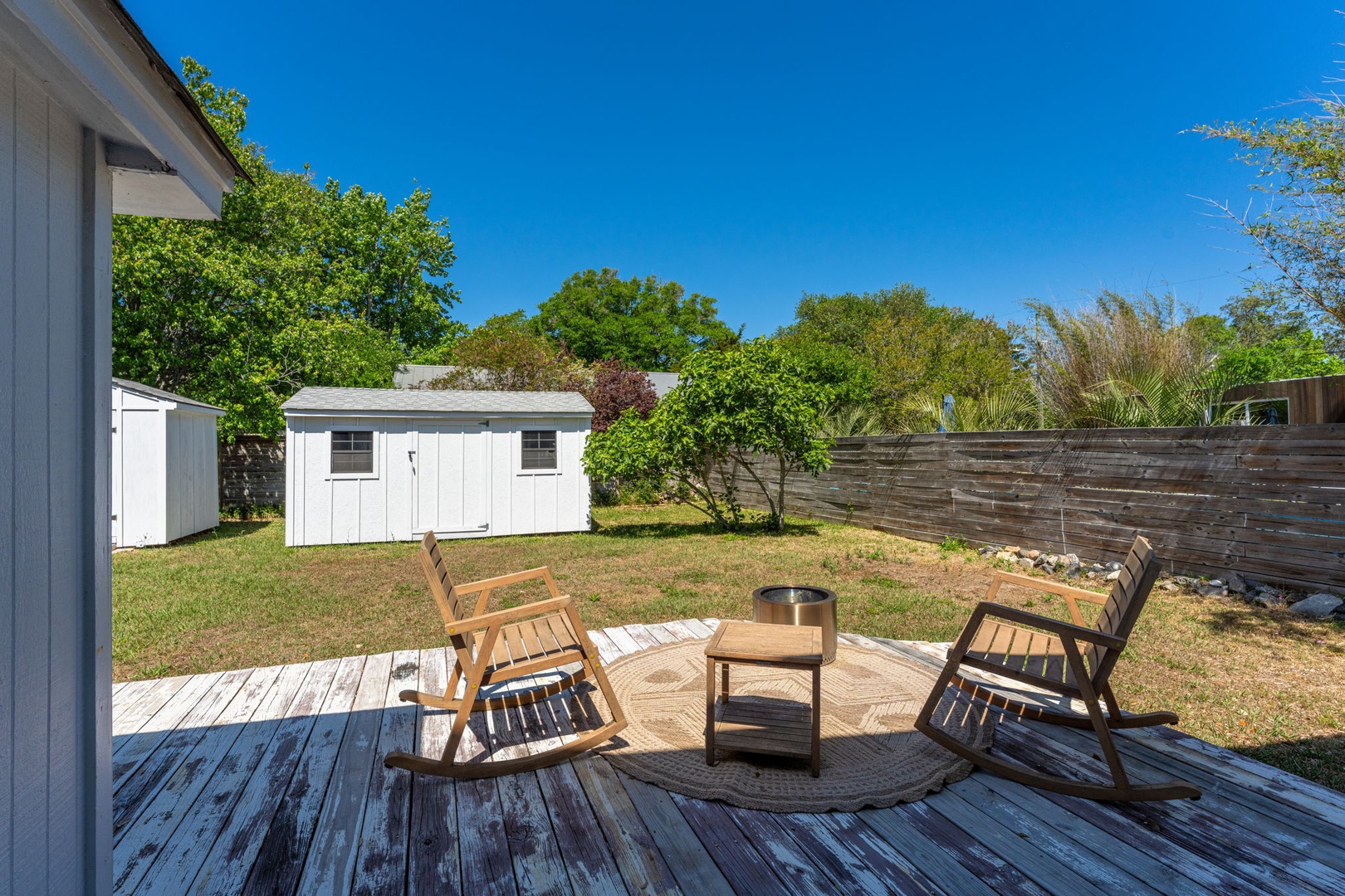 Relax in wooden chairs on your private deck, overlooking a peaceful backyard with lush trees and clear blue skies.