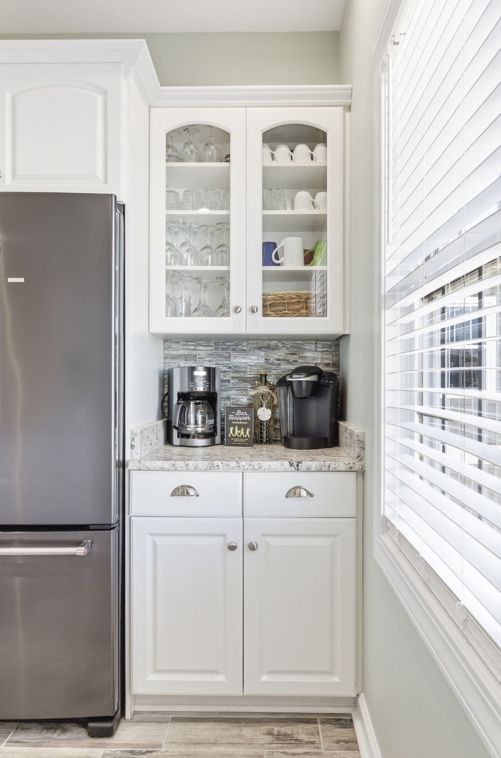 Your personal coffee station awaits, fully stocked with everything you need for that perfect morning brew. Fresh light pours through the window as you reach for your favorite mug from the glass-front cabinet.