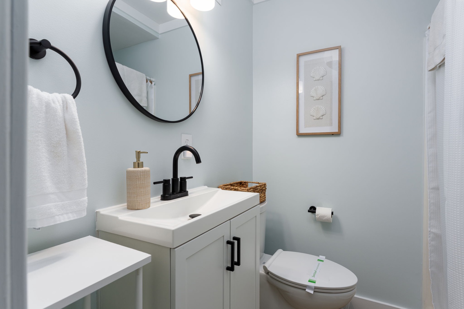 Your private bathroom features a modern vanity with sleek fixtures, a round mirror, and coastal-inspired artwork that creates a serene, spa-like atmosphere for your stay.