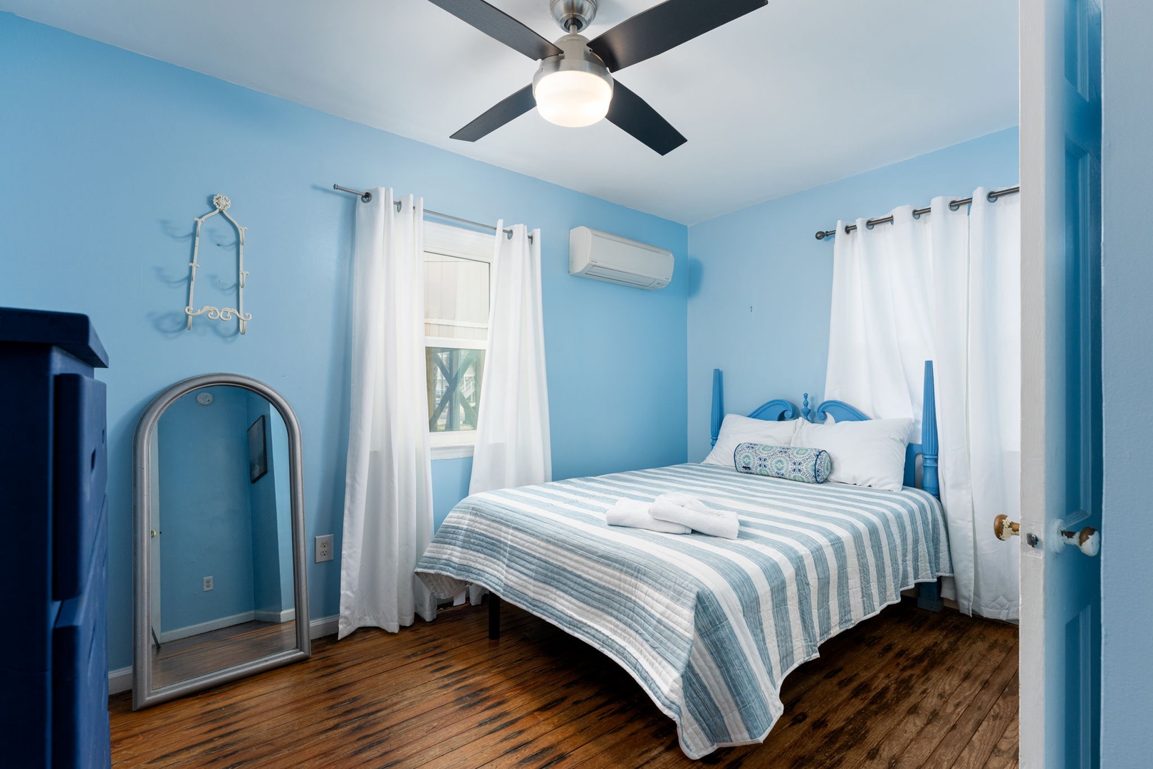 Sink into crisp linens and coastal blues in your serene bedroom retreat. A ceiling fan keeps you cool while white curtains frame tropical glimpses, making this your peaceful escape.