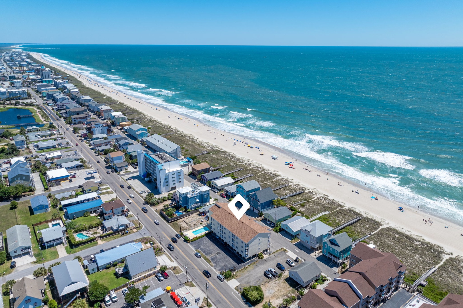 Aerial view of beachfront vacation rentals stretching along pristine coastline, with turquoise waves rolling onto sandy shores and coastal community nearby.