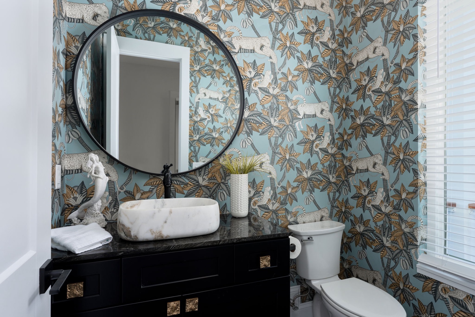 Refresh in your stylish powder room featuring tropical wallpaper, marble vessel sink, and elegant black vanity for a luxurious touch.