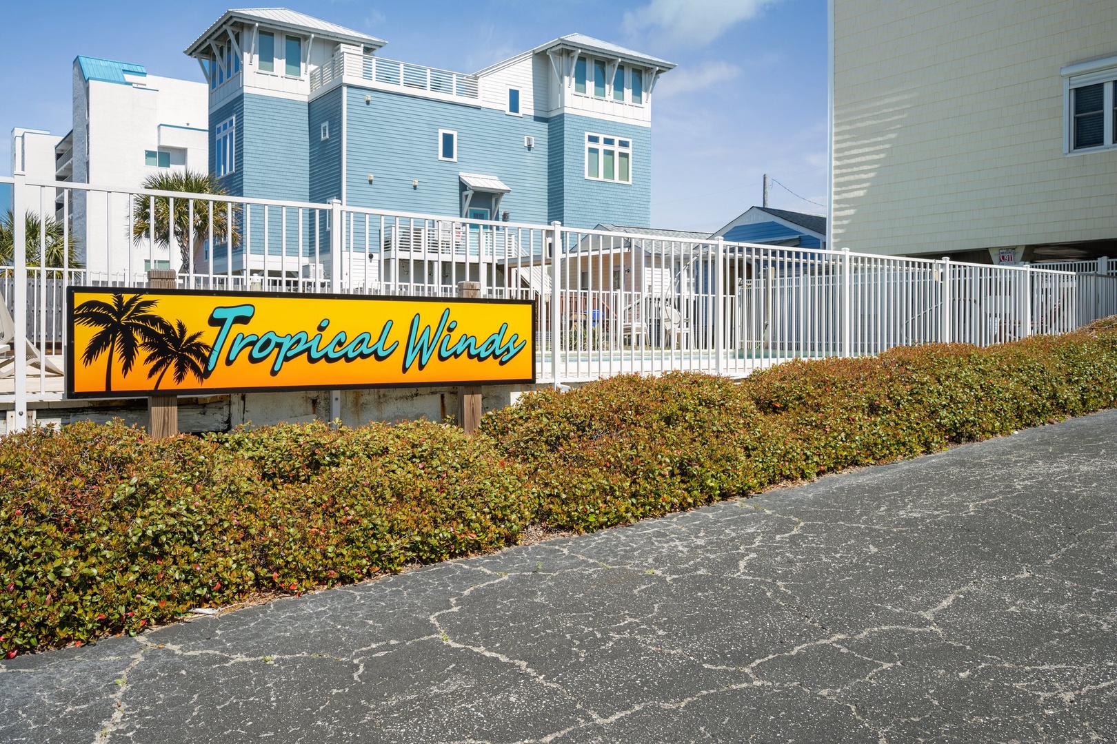 Tropical Winds vacation rental complex entrance with colorful beach house buildings and white fence surrounding the property.