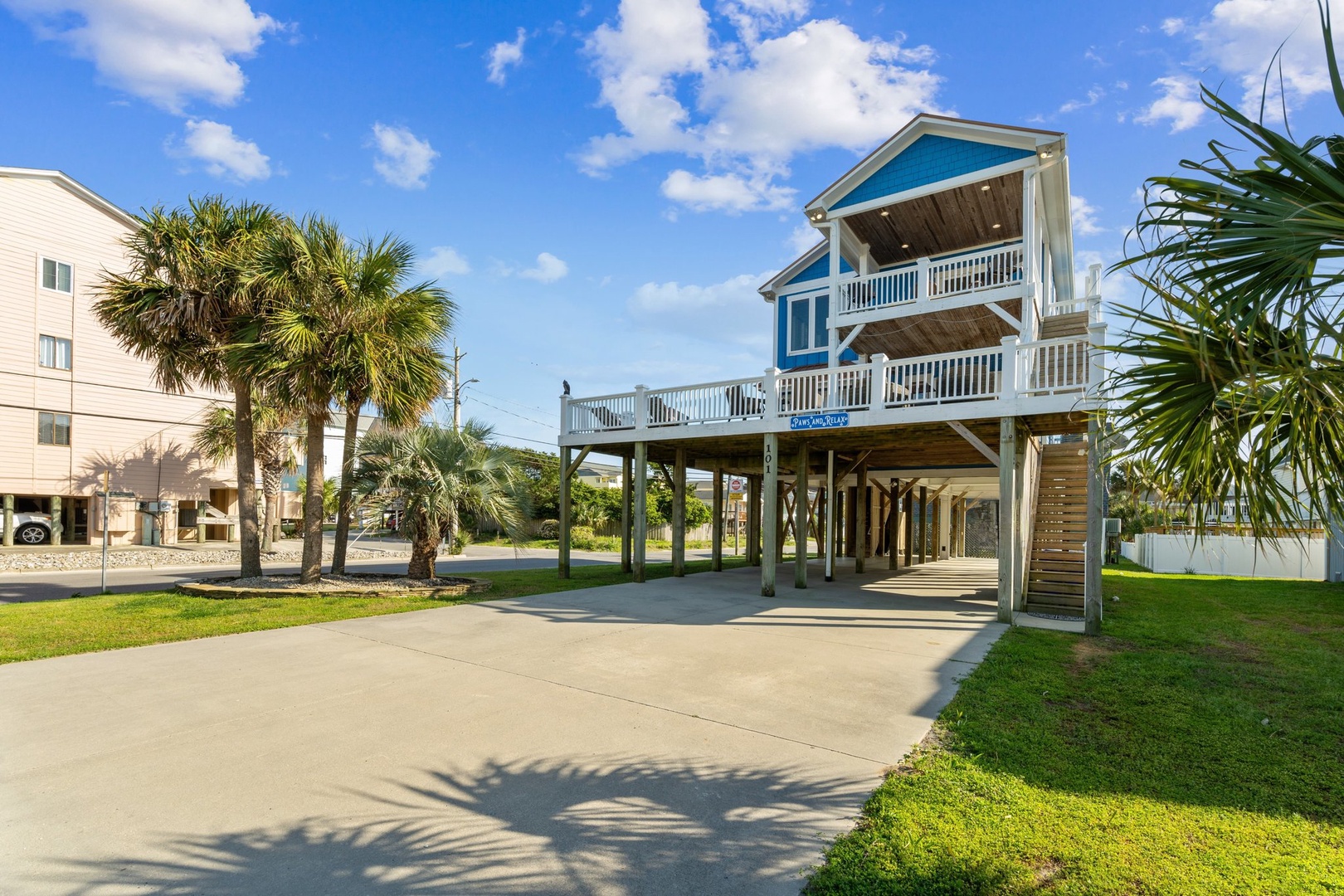 Coastal beach house elevated on stilts with multiple decks and covered parking, surrounded by tropical palm trees.
