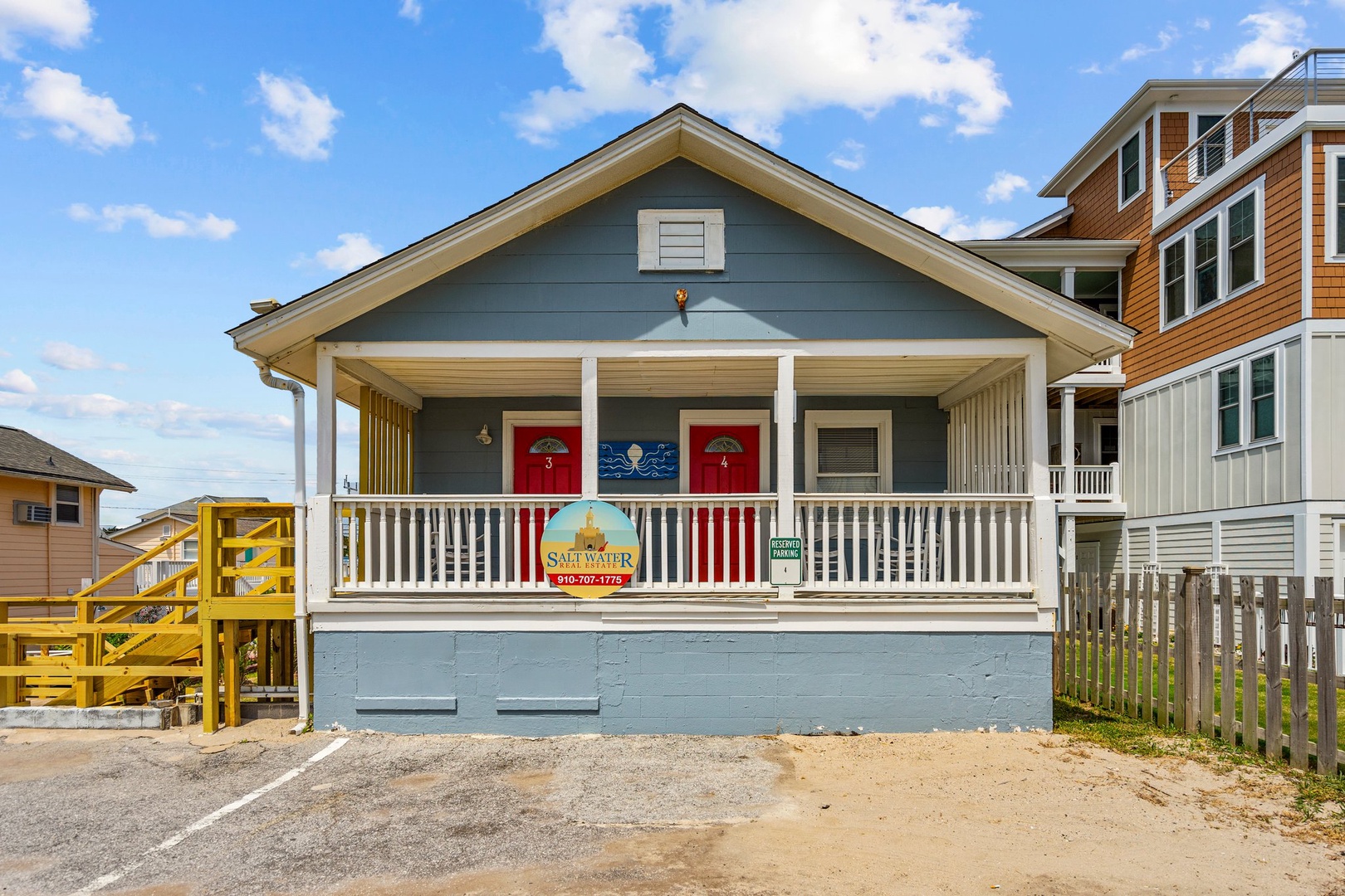 Charming blue beach cottage with covered front porch and distinctive red doors, nestled among colorful coastal homes.