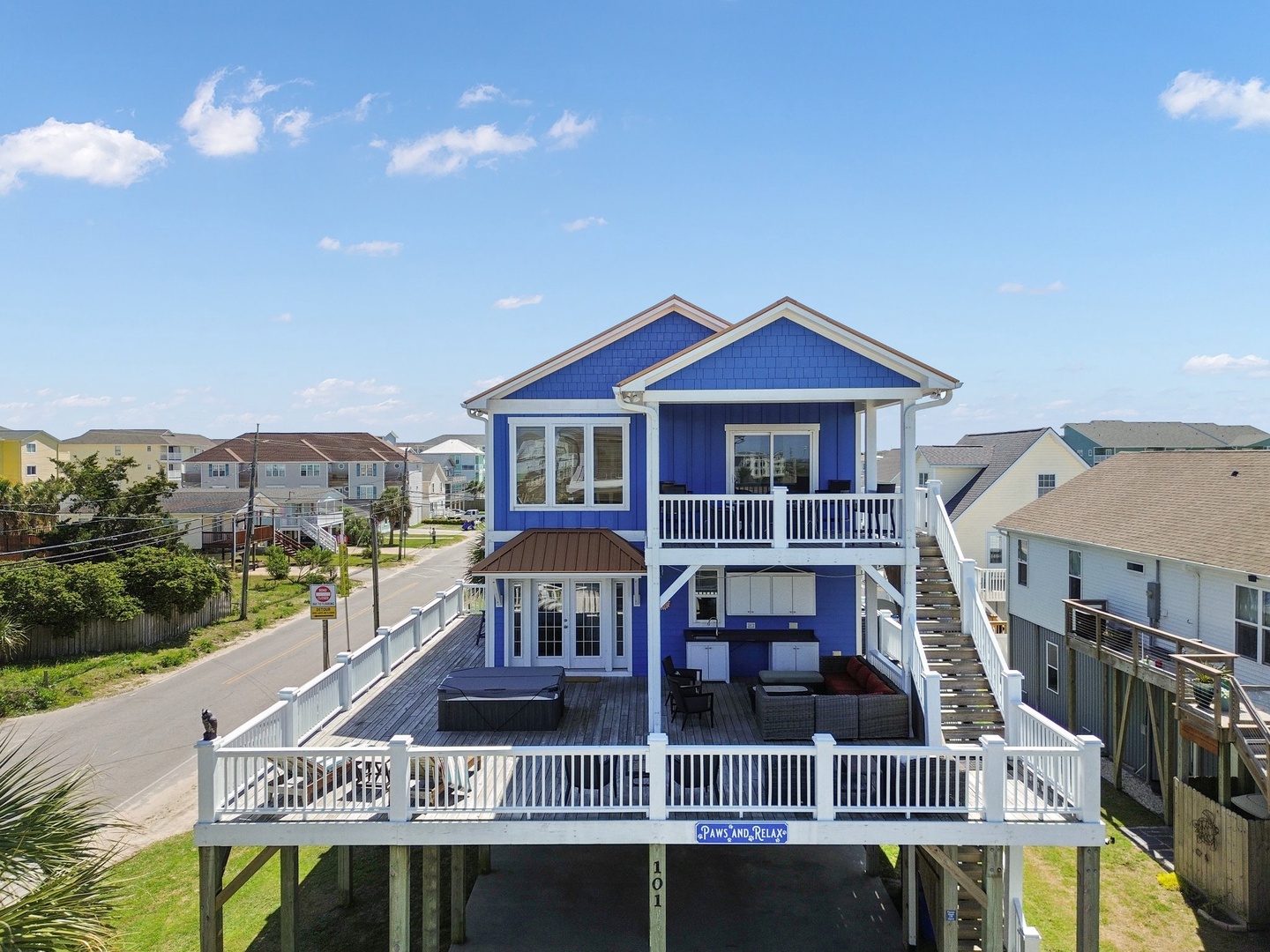 Elevated blue beach house with wraparound decks and stairs, nestled among coastal homes under a clear summer sky.