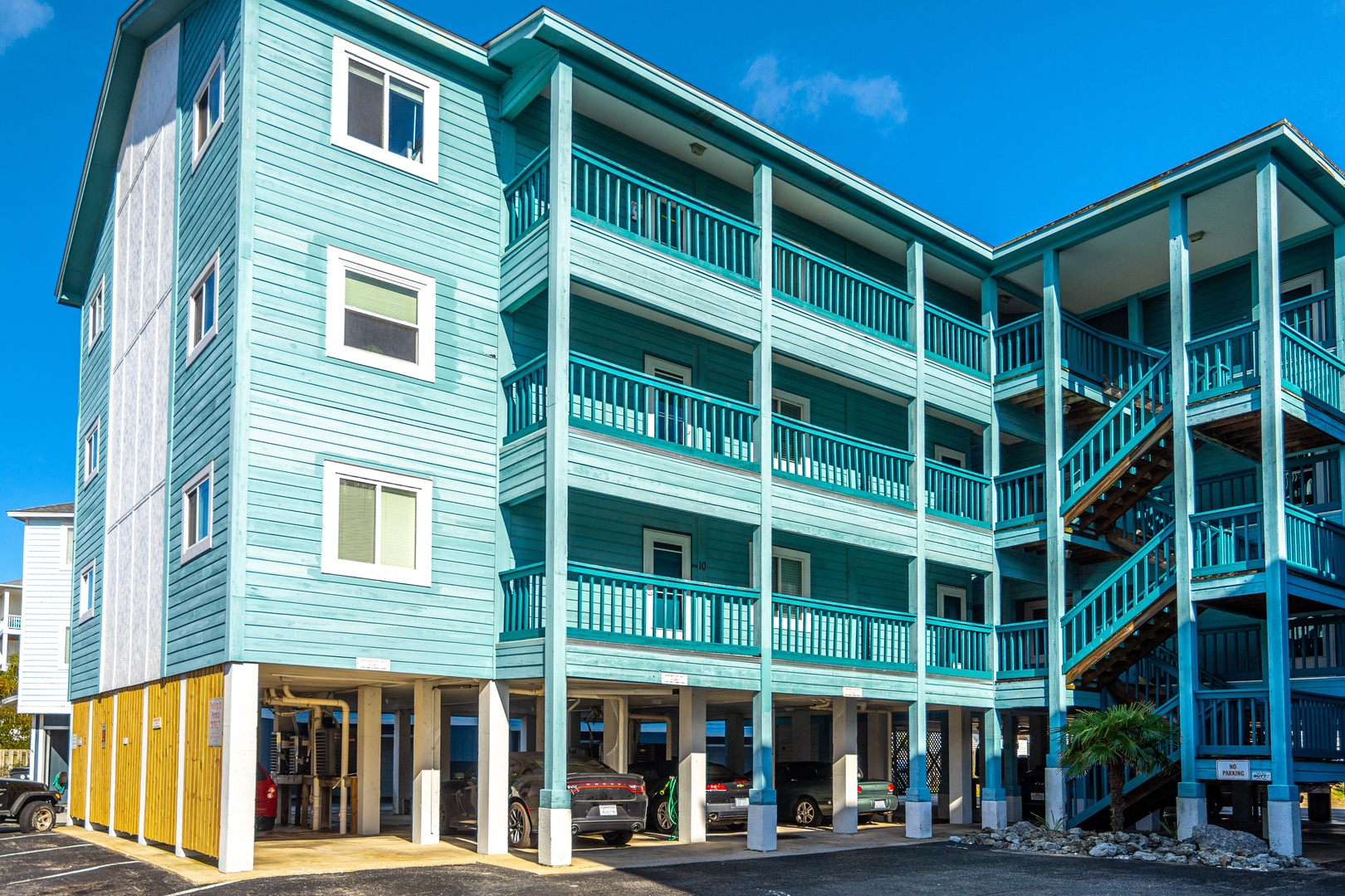 Bright turquoise coastal building featuring covered parking and multi-level balconies under a stunning blue sky.