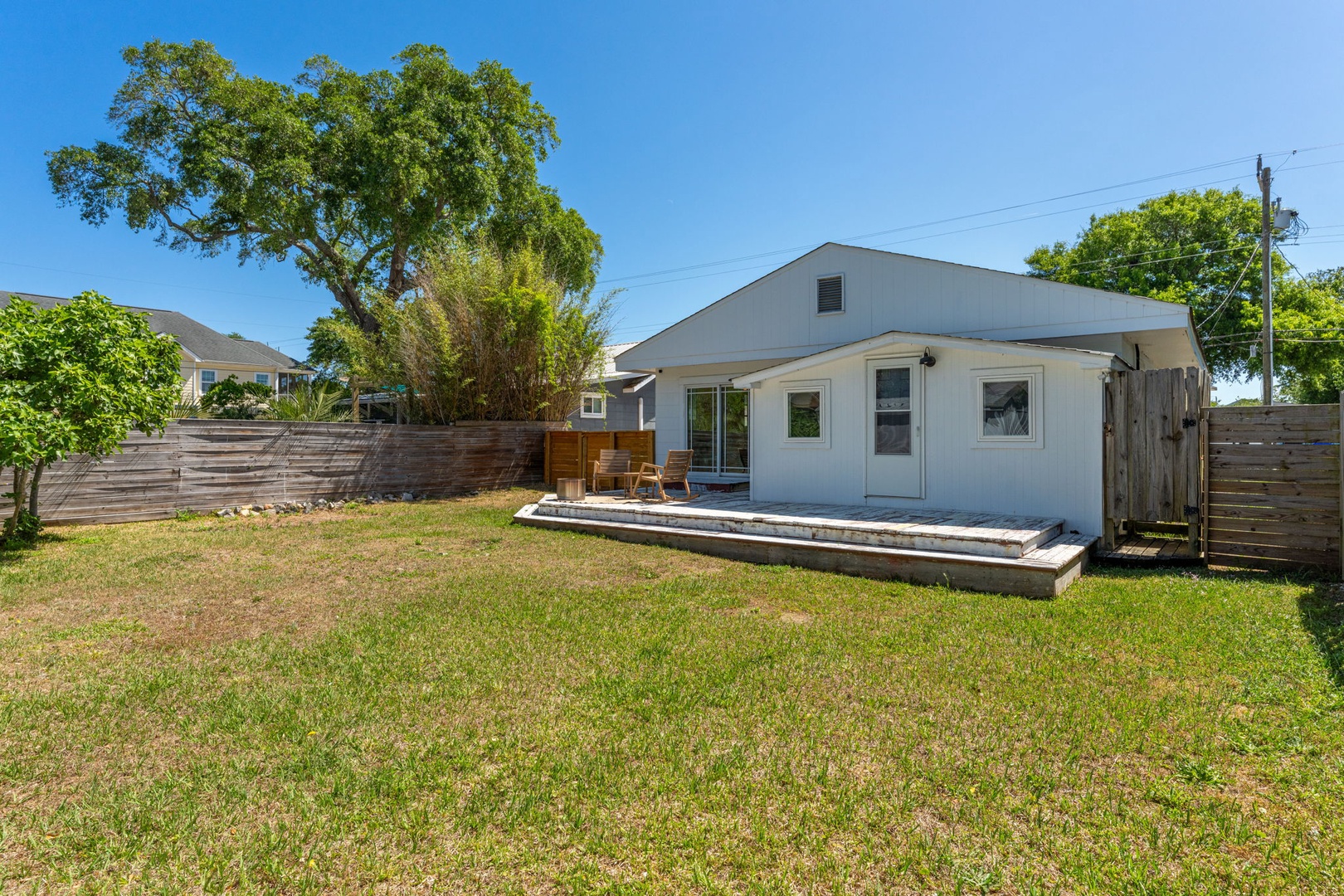 A charming single-story home with spacious backyard featuring mature trees and a private patio deck under bright blue skies.