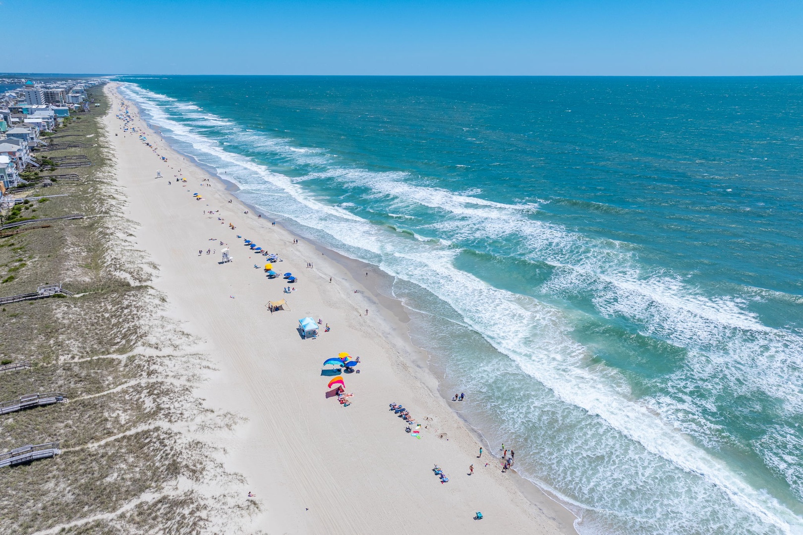 Stunning aerial view of pristine beachfront with turquoise waters and soft white sand stretching along the coastline, dotted with colorful umbrellas and nearby resort buildings.