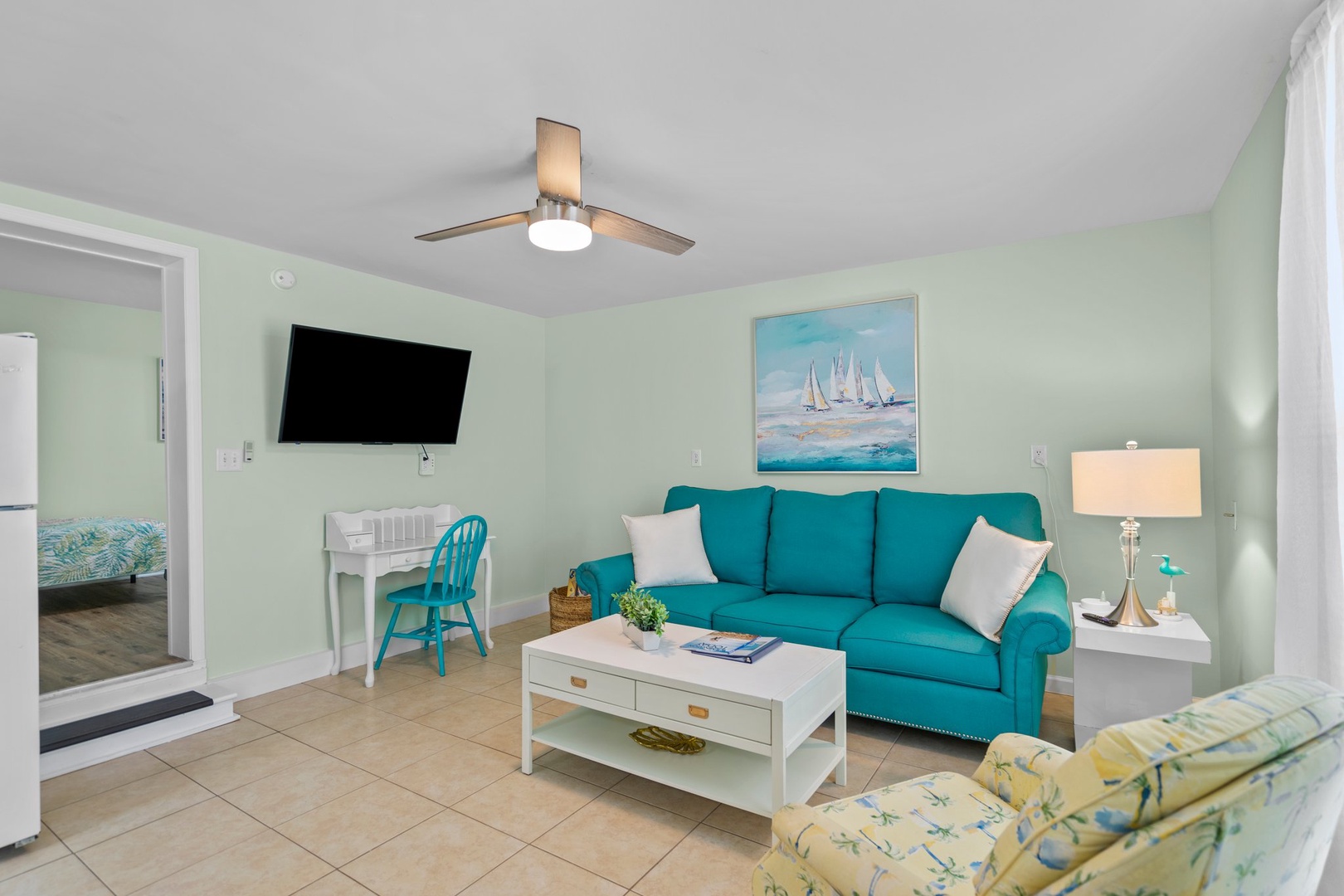 Relax in your coastal living room with vibrant teal sofa, nautical artwork, and ceiling fan keeping you comfortable.