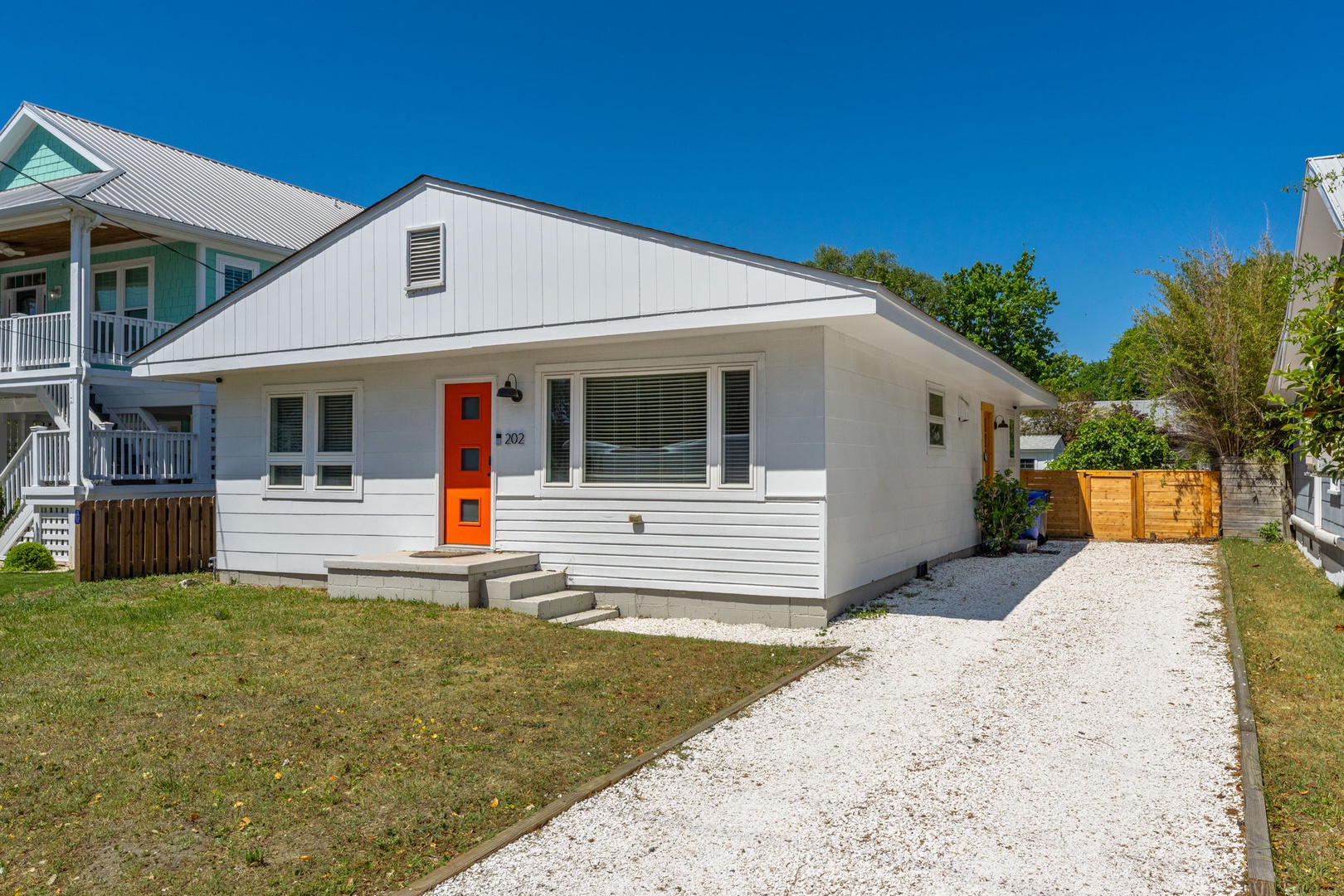 Modern beach cottage with vibrant orange door and gravel driveway, nestled in a charming coastal neighborhood.