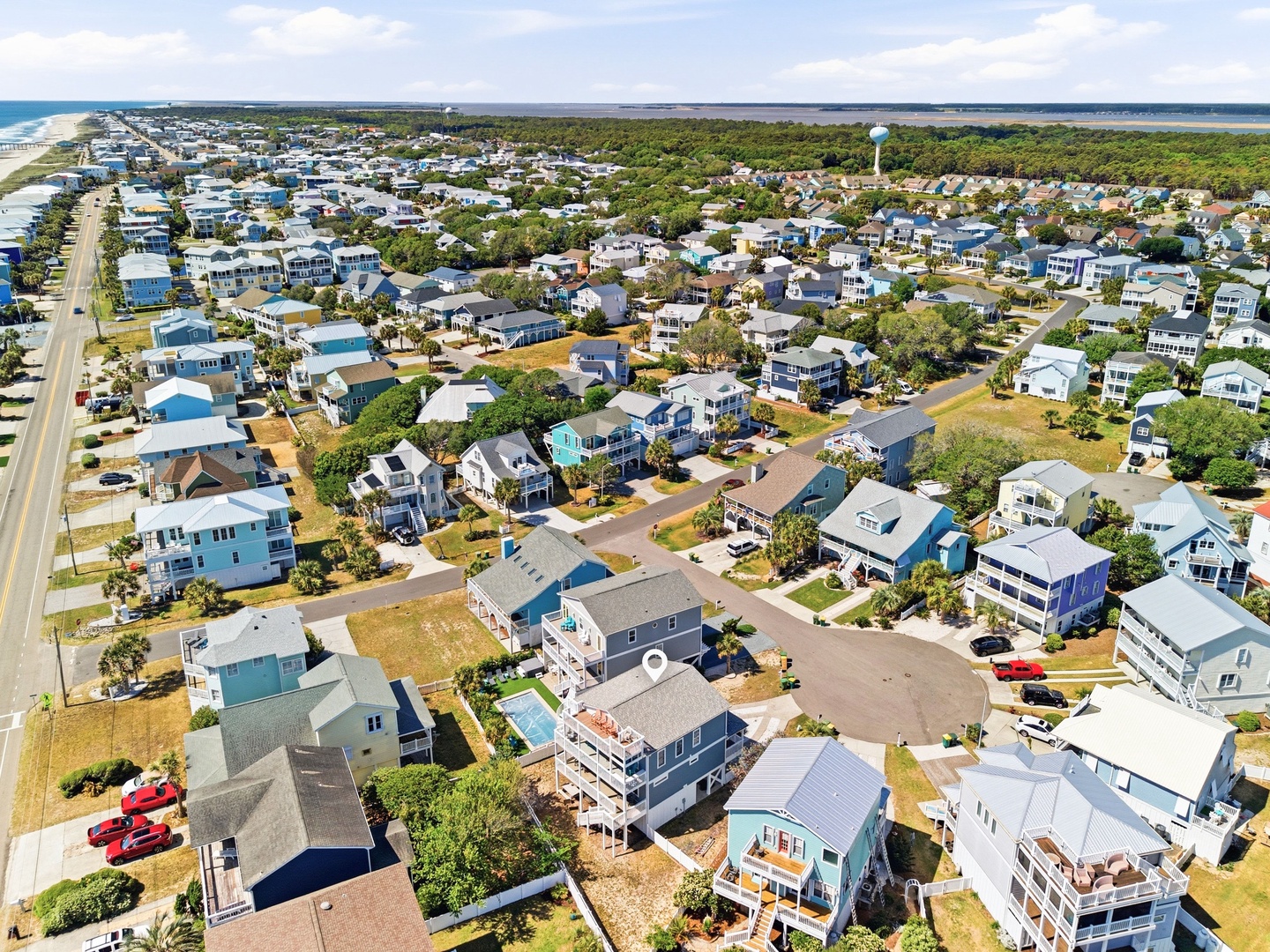 Aerial view of a charming coastal neighborhood showcasing beachside vacation rental properties nestled between the ocean and inland waterways, surrounded by lush greenery and tree-lined streets.