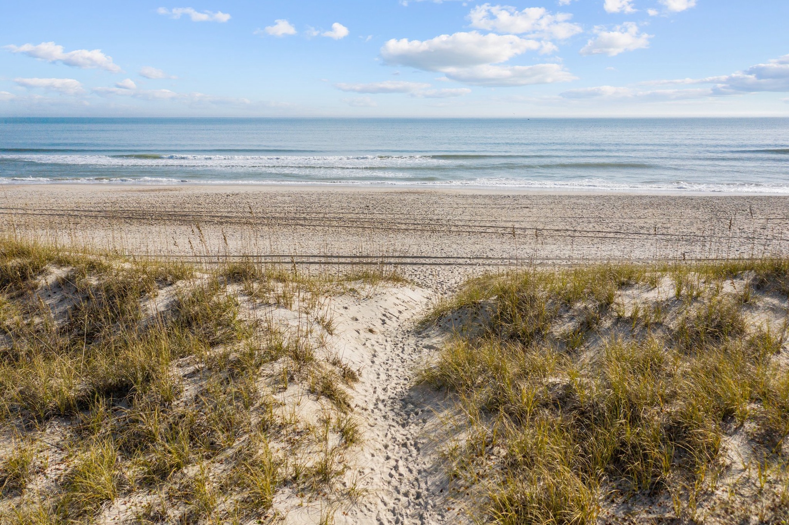 Pristine sandy beach with natural dunes and gentle waves under a beautiful blue sky with scattered white clouds.