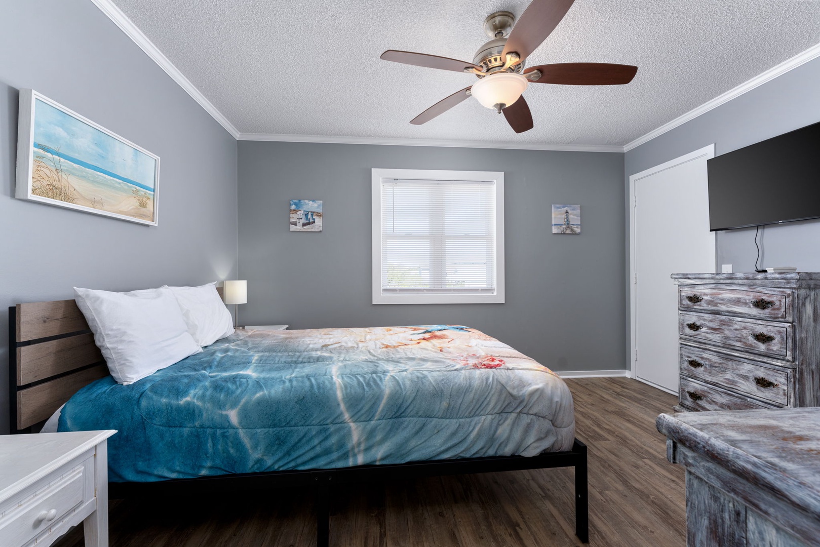 Drift off to sleep in your calming coastal bedroom, complete with a plush bed, ceiling fan, and beach-inspired artwork. The weathered wood dresser and mounted TV add comfort and convenience.