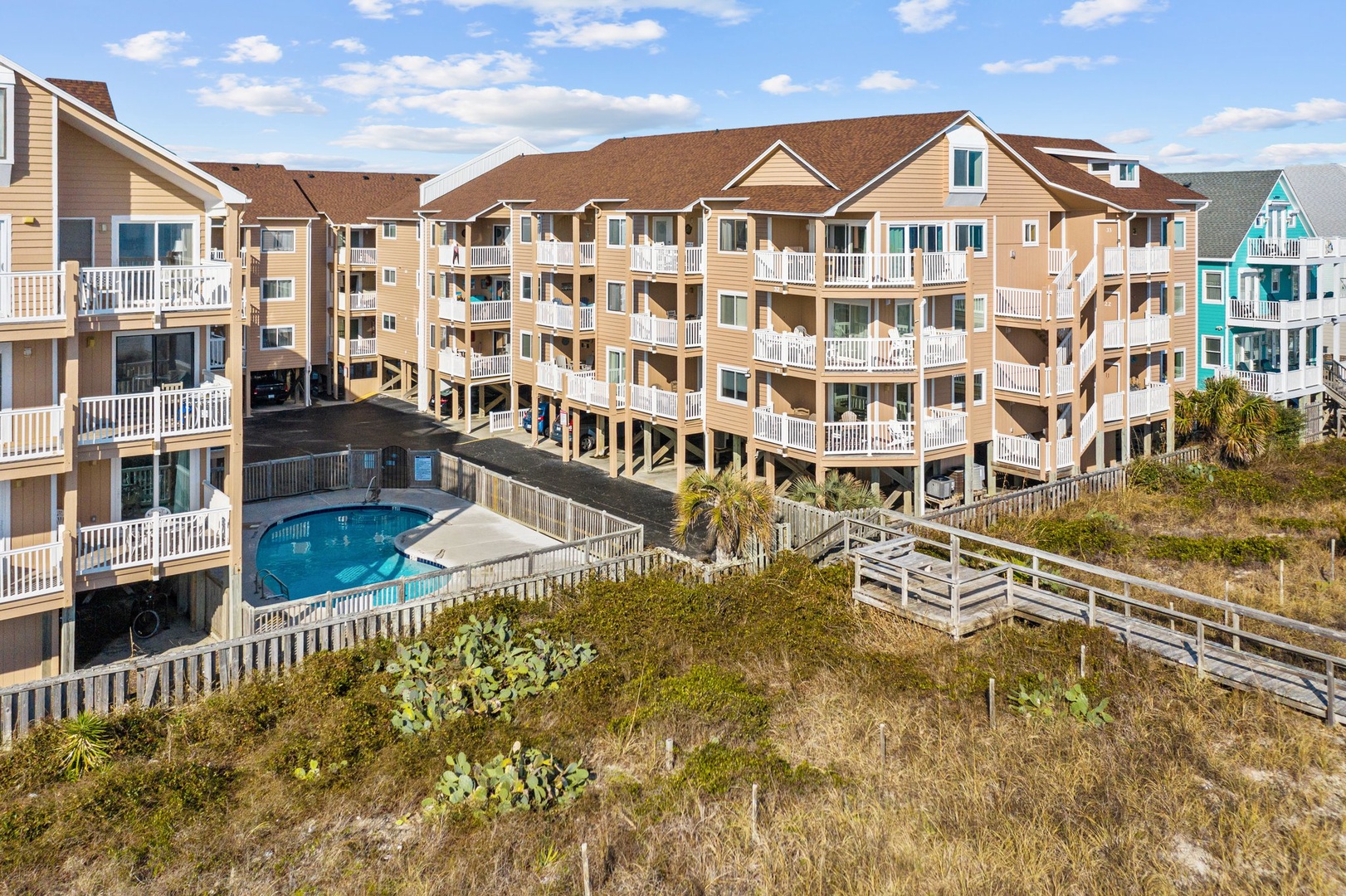Coastal vacation complex with swimming pool and beach access walkway, featuring multi-level accommodations surrounded by natural dune vegetation.