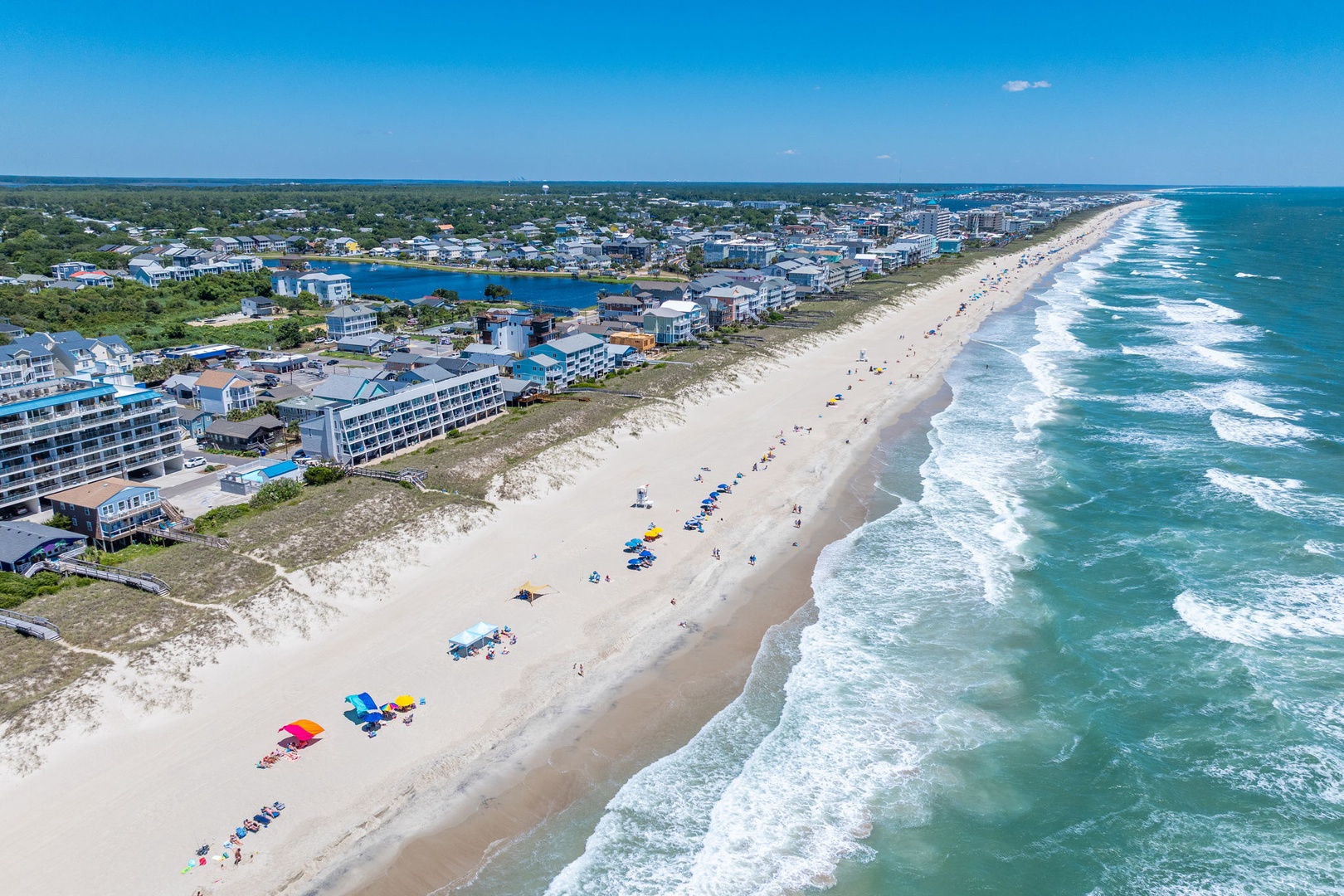 Aerial view of a pristine coastal area featuring a wide sandy beach, rolling ocean waves, and beachfront development. The surrounding neighborhood includes residential areas and waterfront access.