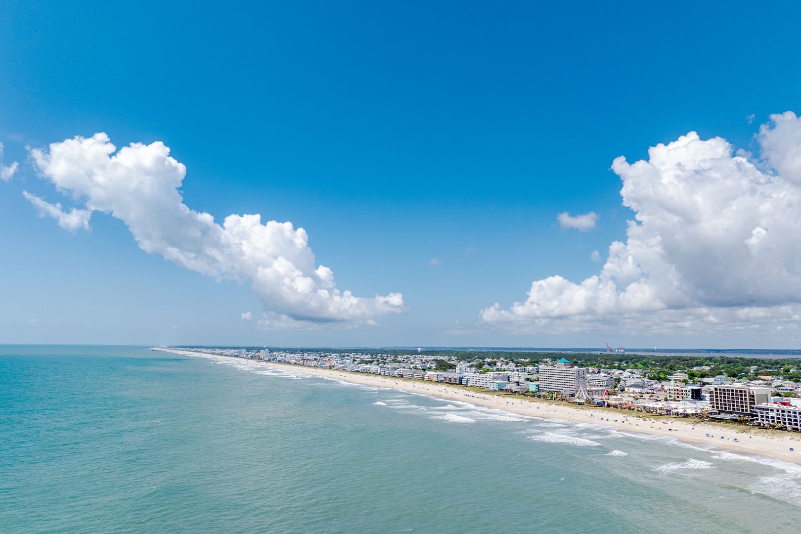 Aerial view captures the property's coastal location along a pristine sandy beach, with calm turquoise waters and a vibrant beachfront community stretching into the distance under a bright sky.