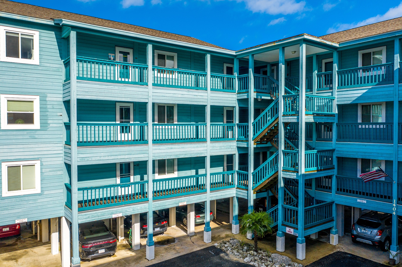 Charming turquoise beach-style building featuring multiple levels of covered parking and guest access stairs under a brilliant blue sky.