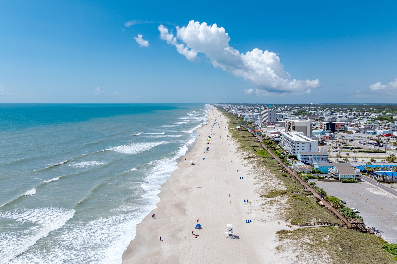 Aerial view of a pristine beach stretching along a coastal town, with soft white sand meeting gentle waves and a scenic boardwalk in the surrounding area.