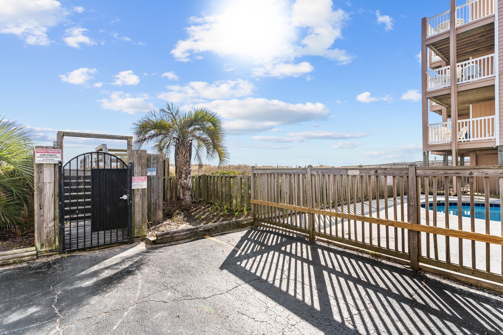 Private gated entrance leads to your coastal retreat with palm trees and pool access under sunny skies.
