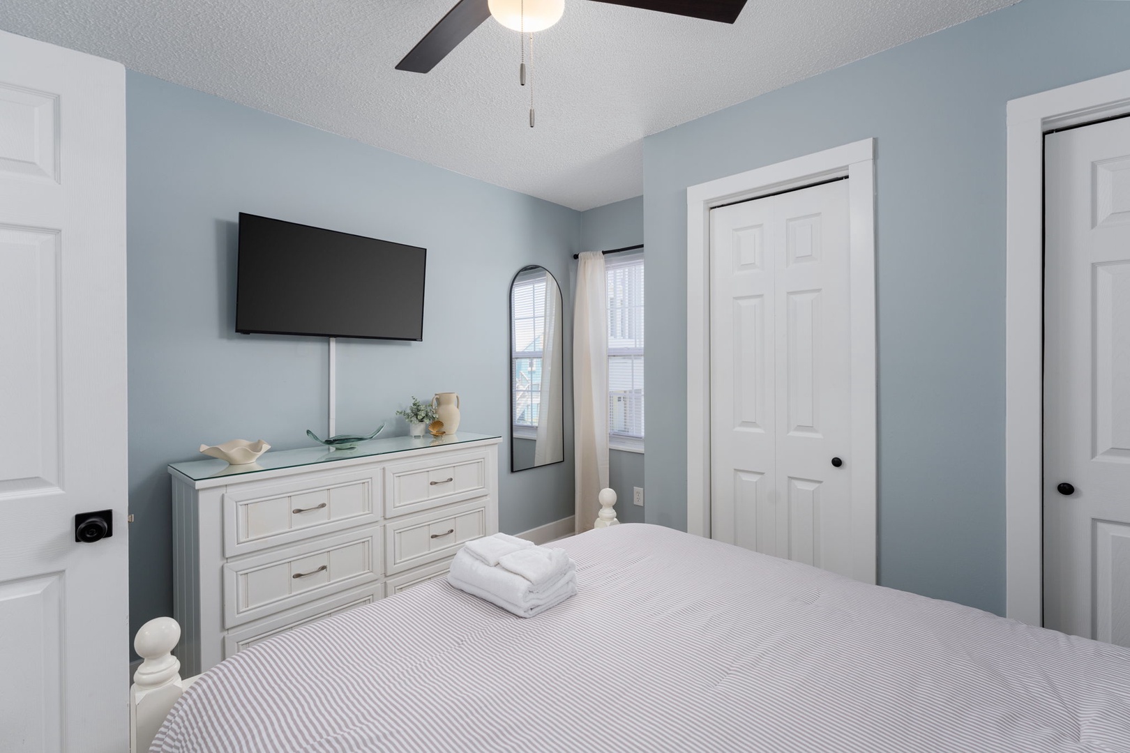 Unwind in your serene bedroom featuring soft blue tones, a plush bed, ceiling fan, flat-screen TV, and ample storage—your personal retreat awaits.