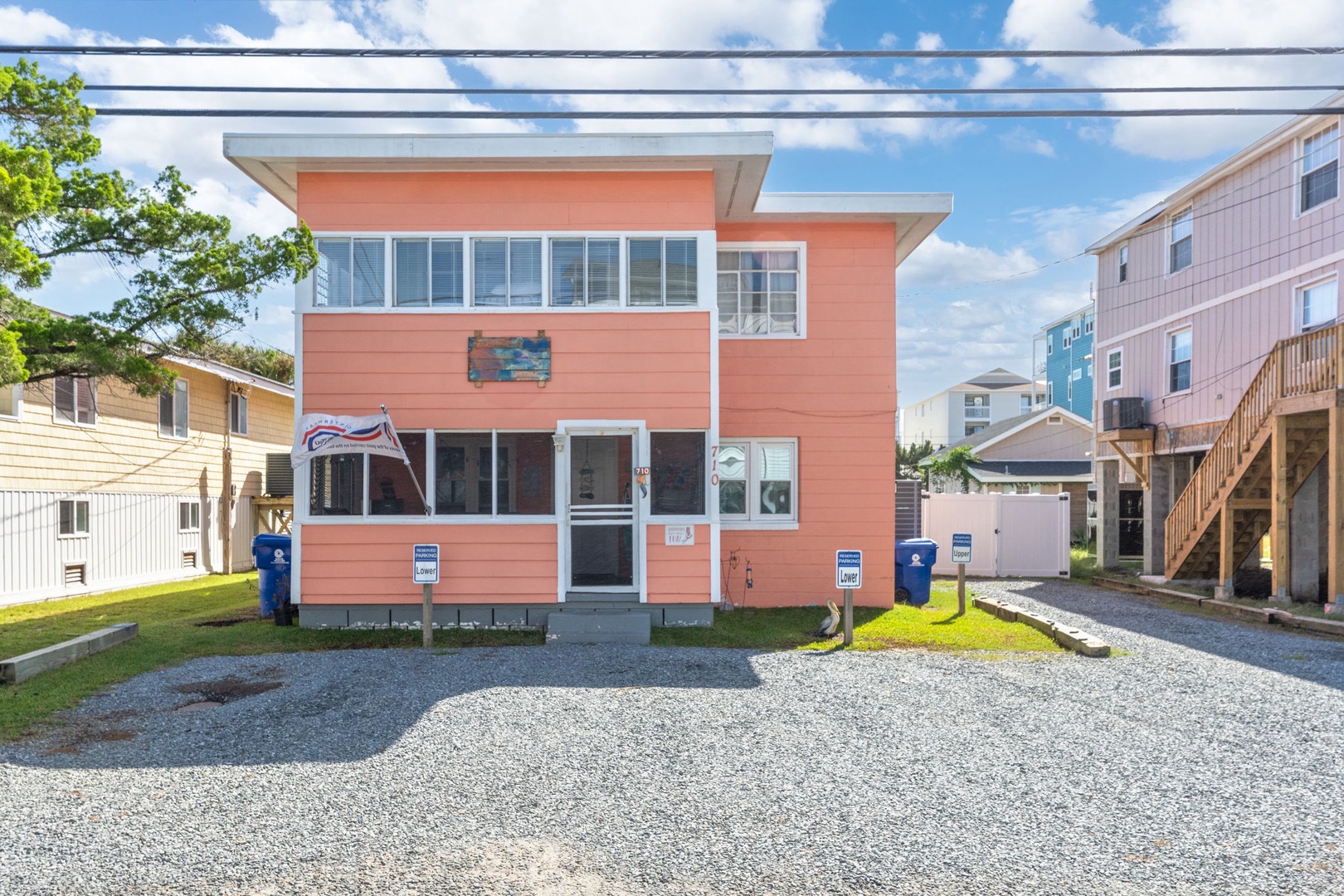 Bright coral-colored beach house with modern design sits in a coastal neighborhood, featuring convenient gravel parking and surrounded by other vacation homes.