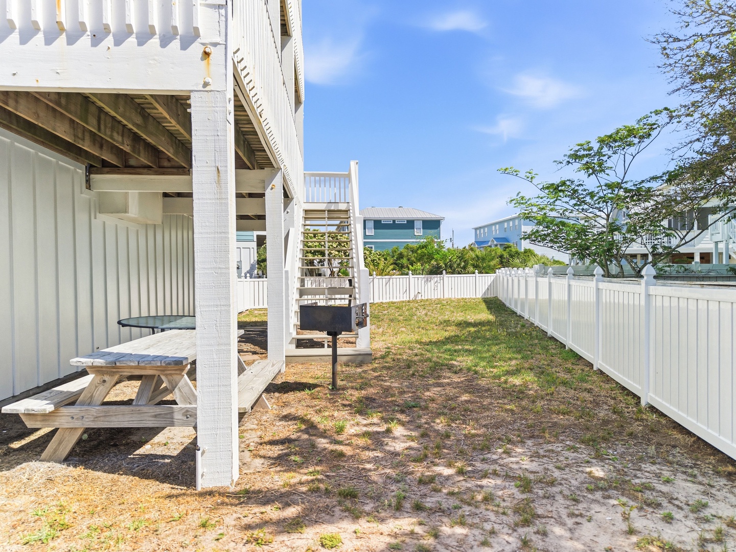 A spacious fenced yard provides a private outdoor retreat beneath the elevated beach house, featuring a picnic table and convenient grill area for relaxed coastal living.