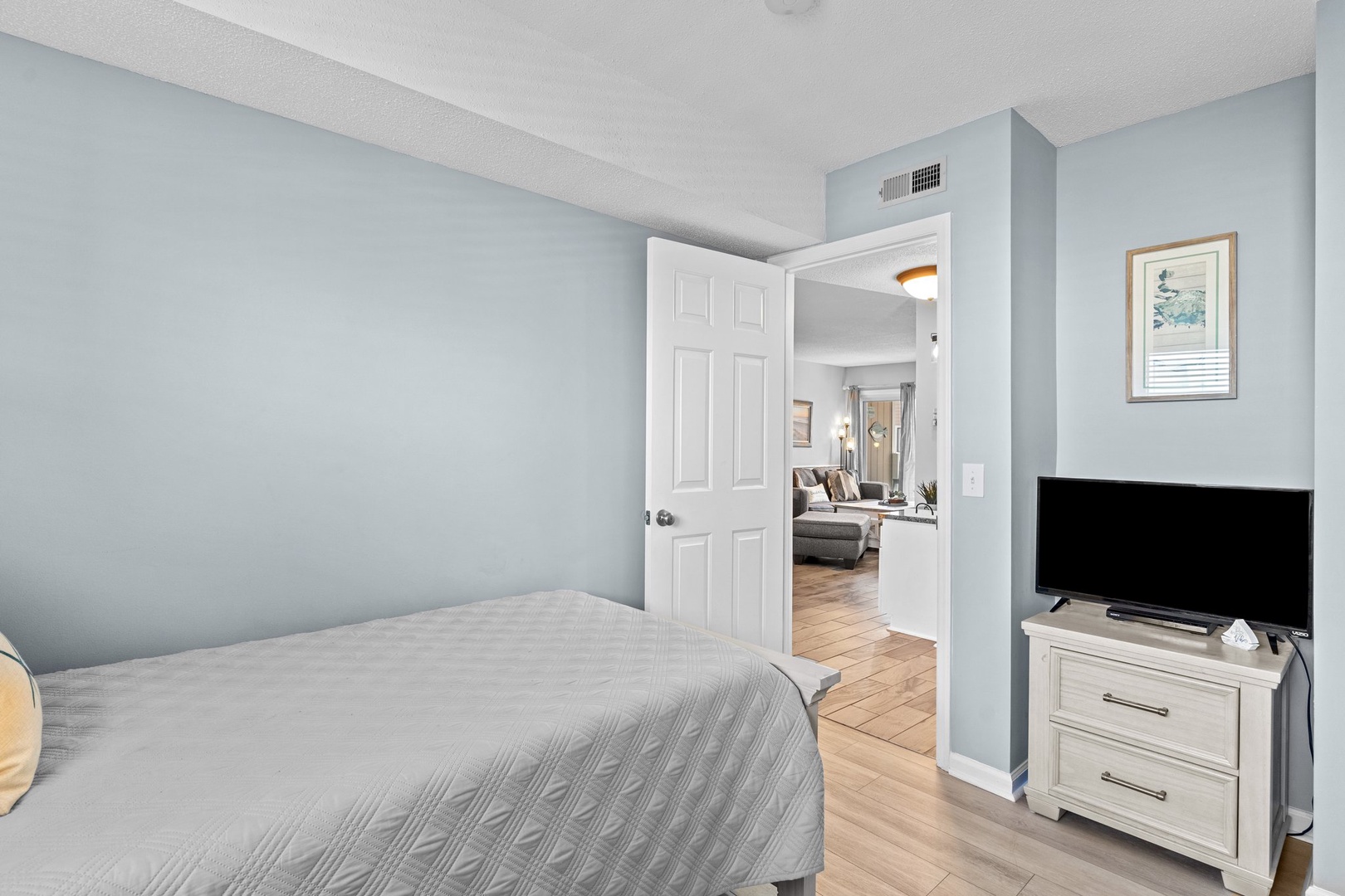 Your peaceful bedroom features soft blue walls and connects to a bright living area through an open doorway.