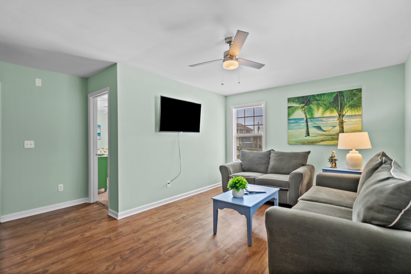 Unwind in your coastal-themed living room with soft mint walls, comfortable seating, and tropical artwork creating a peaceful retreat.