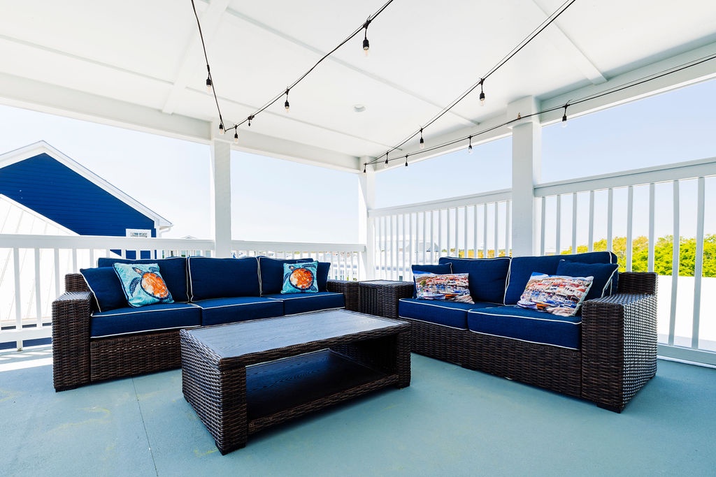 Sink into plush navy cushions on your covered deck, where string lights create evening magic and coastal breezes flow through.