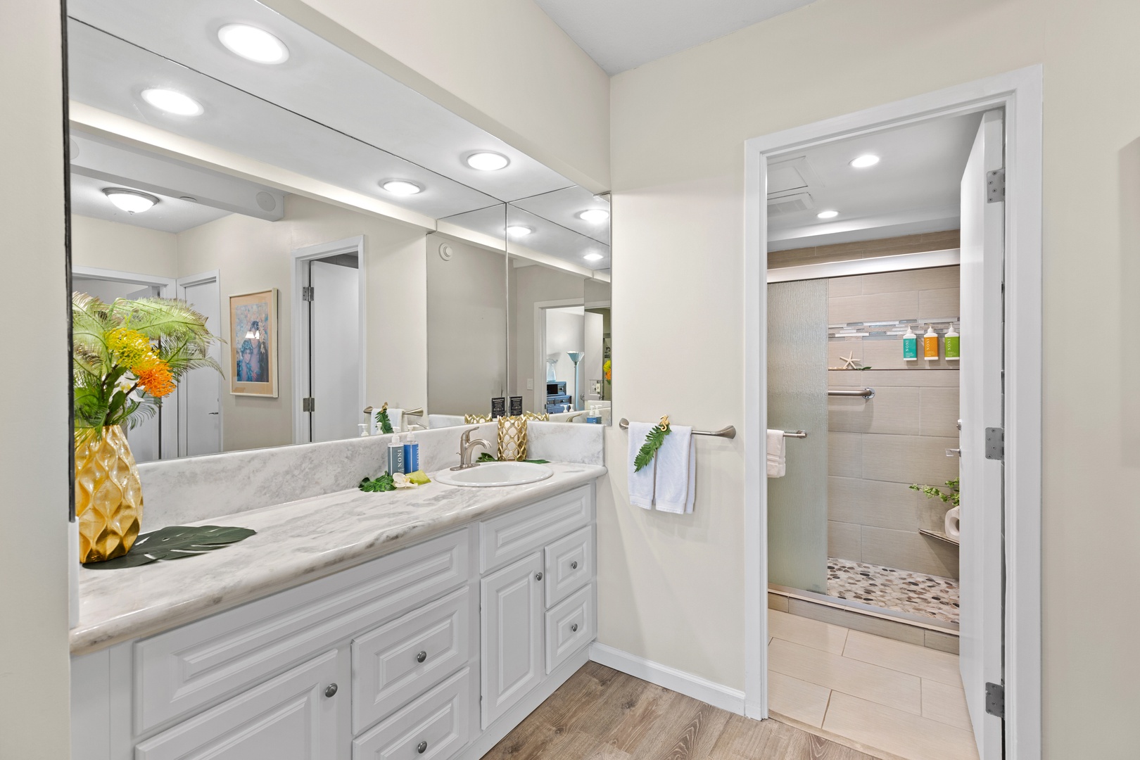 Your private bathroom offers bright lighting, marble counters, fresh florals, and a beautifully tiled walk-in shower with thoughtful shelf space and beachy accents.