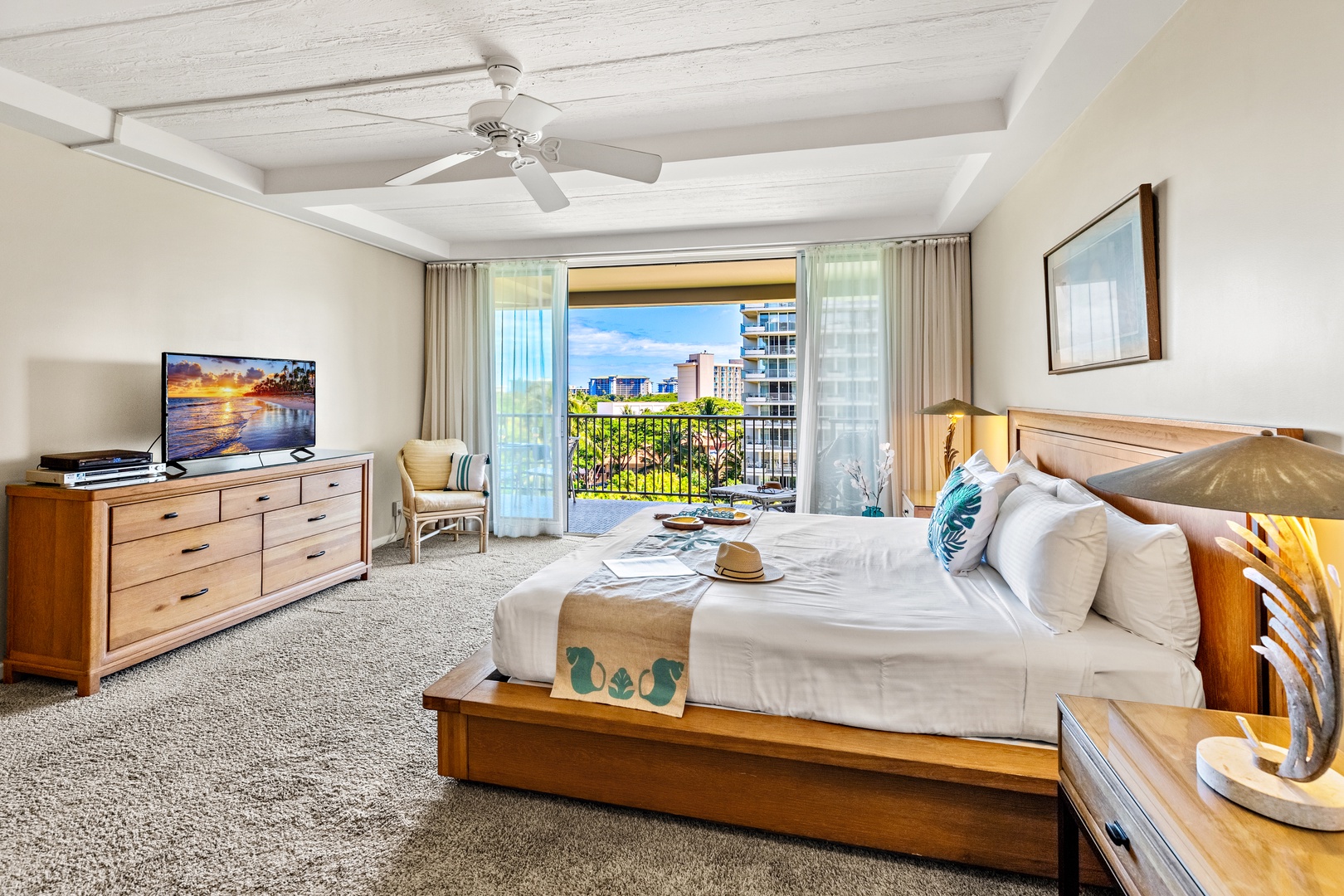 Your spacious bedroom invites tropical relaxation with a breezy ceiling fan overhead and a private balcony framing views of lush greenery and nearby buildings—perfect for morning coffee or sunset moments.