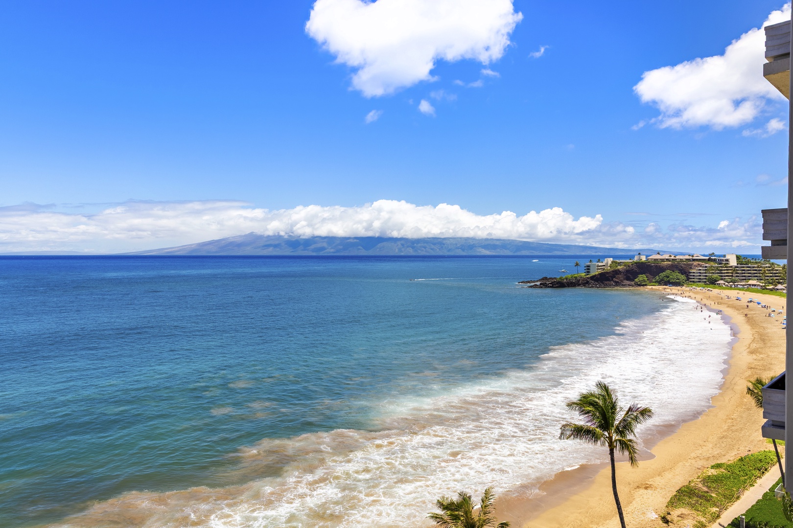 Panoramic coastal view showcasing a pristine sandy beach, turquoise ocean waters, distant mountain ranges, and nearby resort properties under a bright blue sky with scattered clouds.